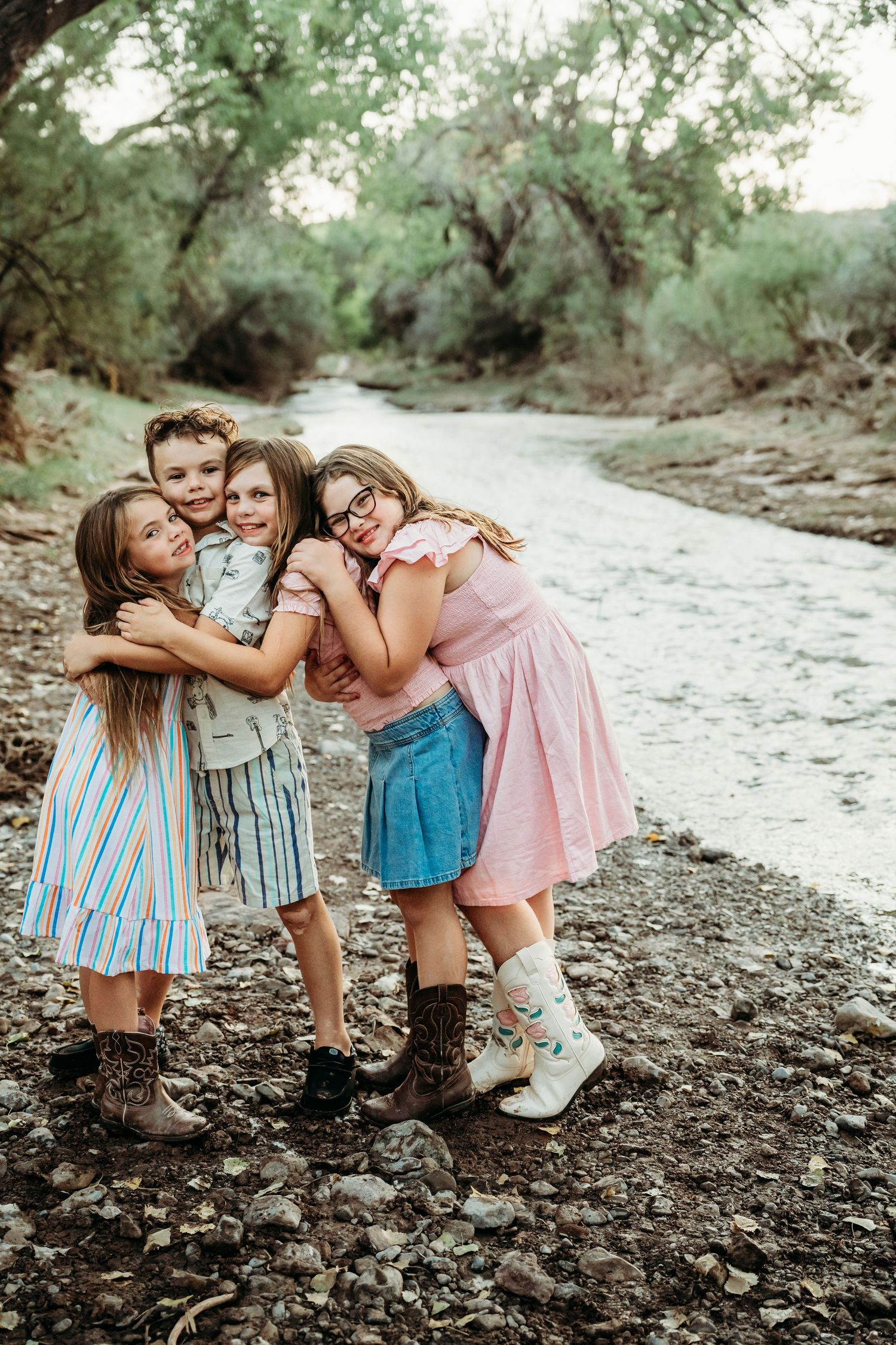 Four children hugging and smiling together on a rocky riverbank surrounded by lush green trees.
