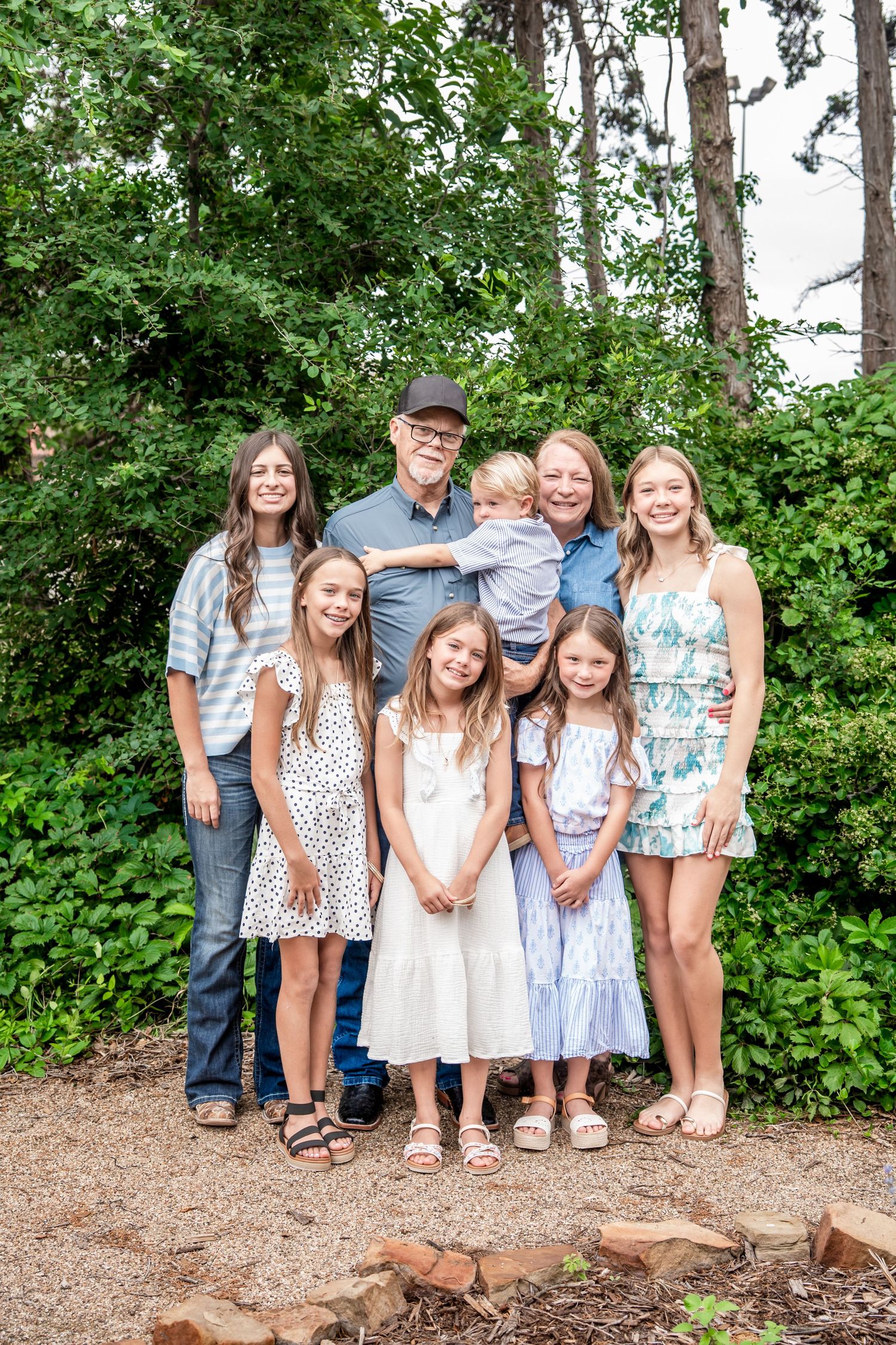 Grandparents pose with six grandchildren of various ages in a lush green outdoor garden setting.