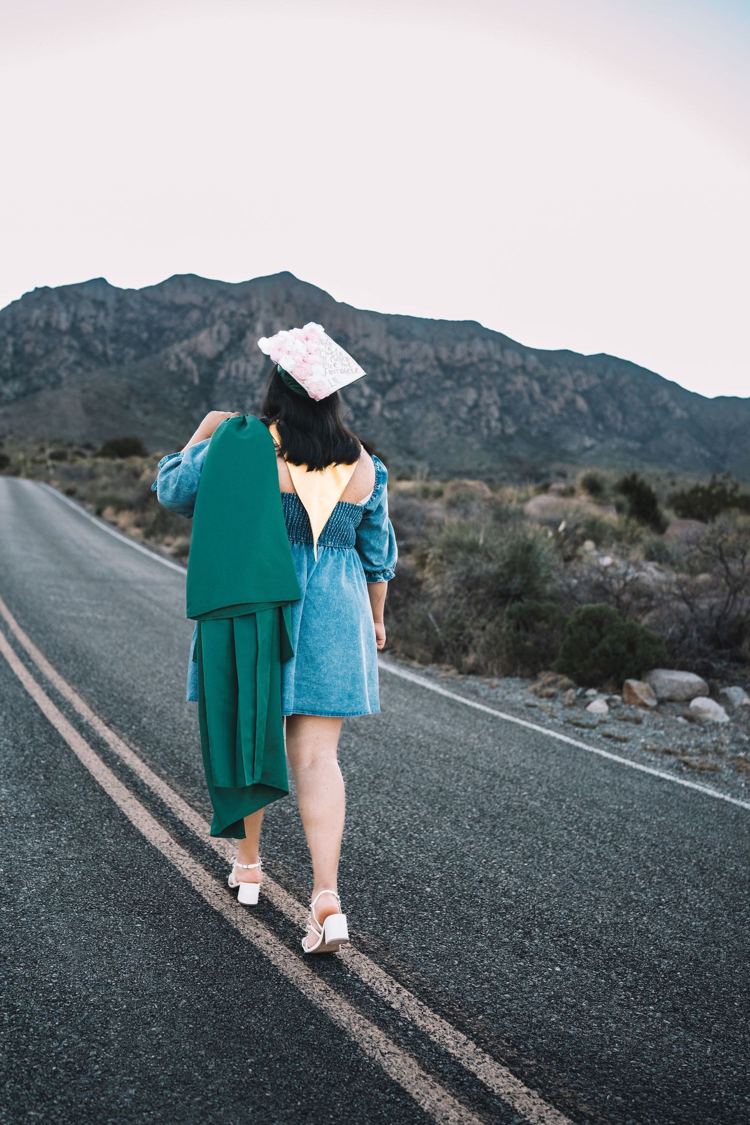 Graduate walking away on a desert road carrying a green graduation gown and decorated cap with mountains ahead.