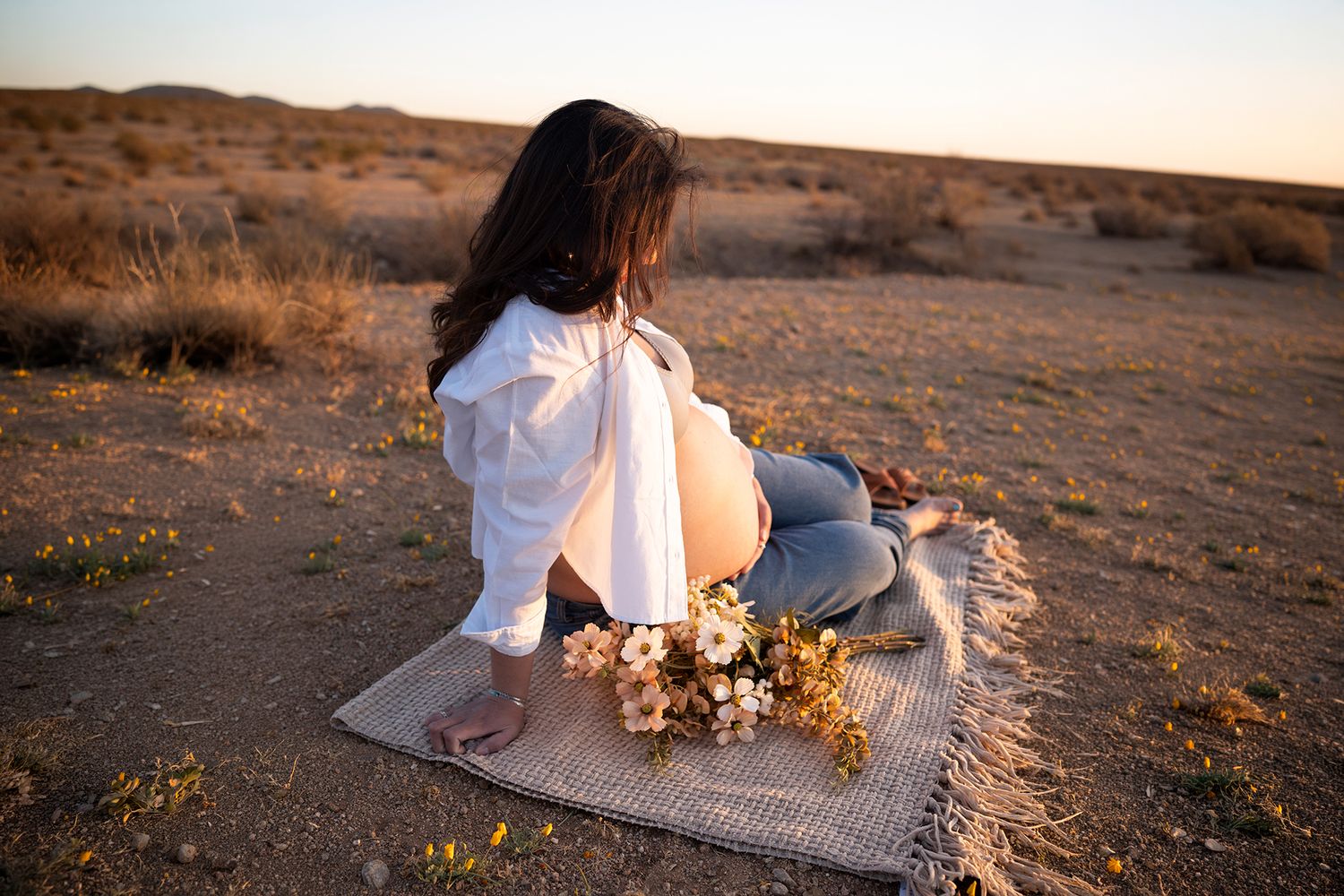 Pregnant woman sitting barefoot on mat in arid desert landscape at sunset with flowers.