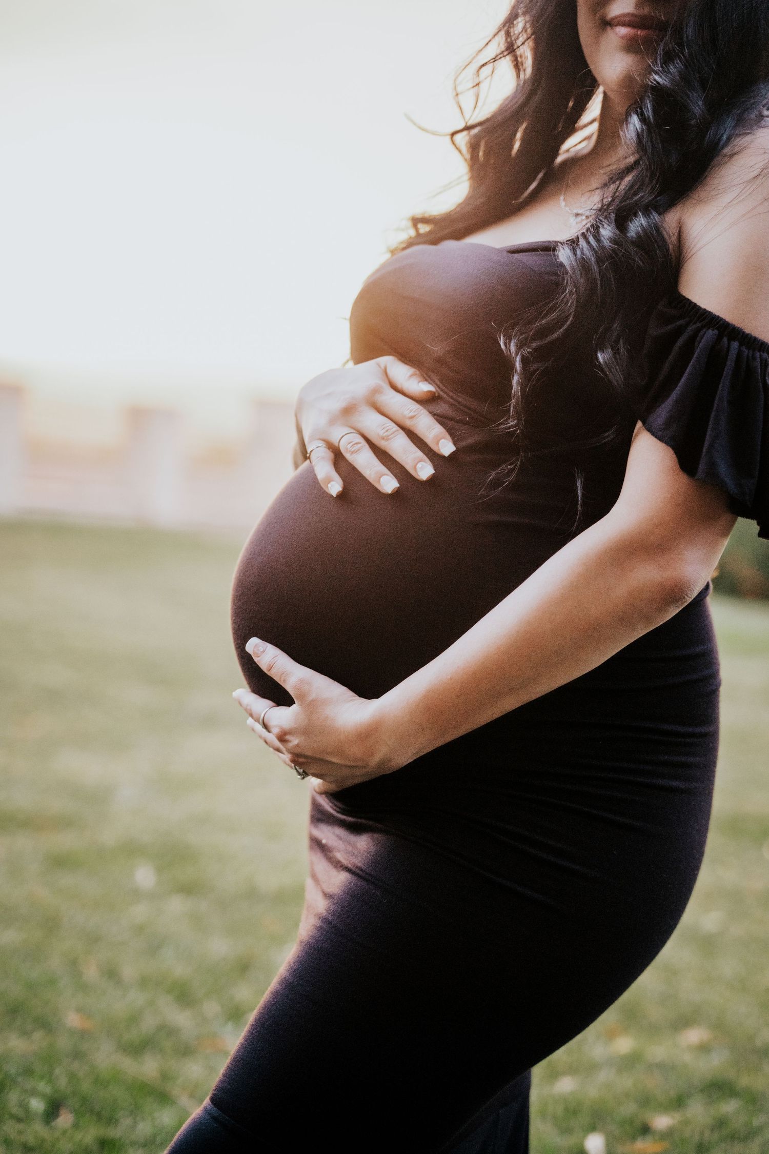 Pregnant woman with tattoos in black off-shoulder dress gently holding baby bump.