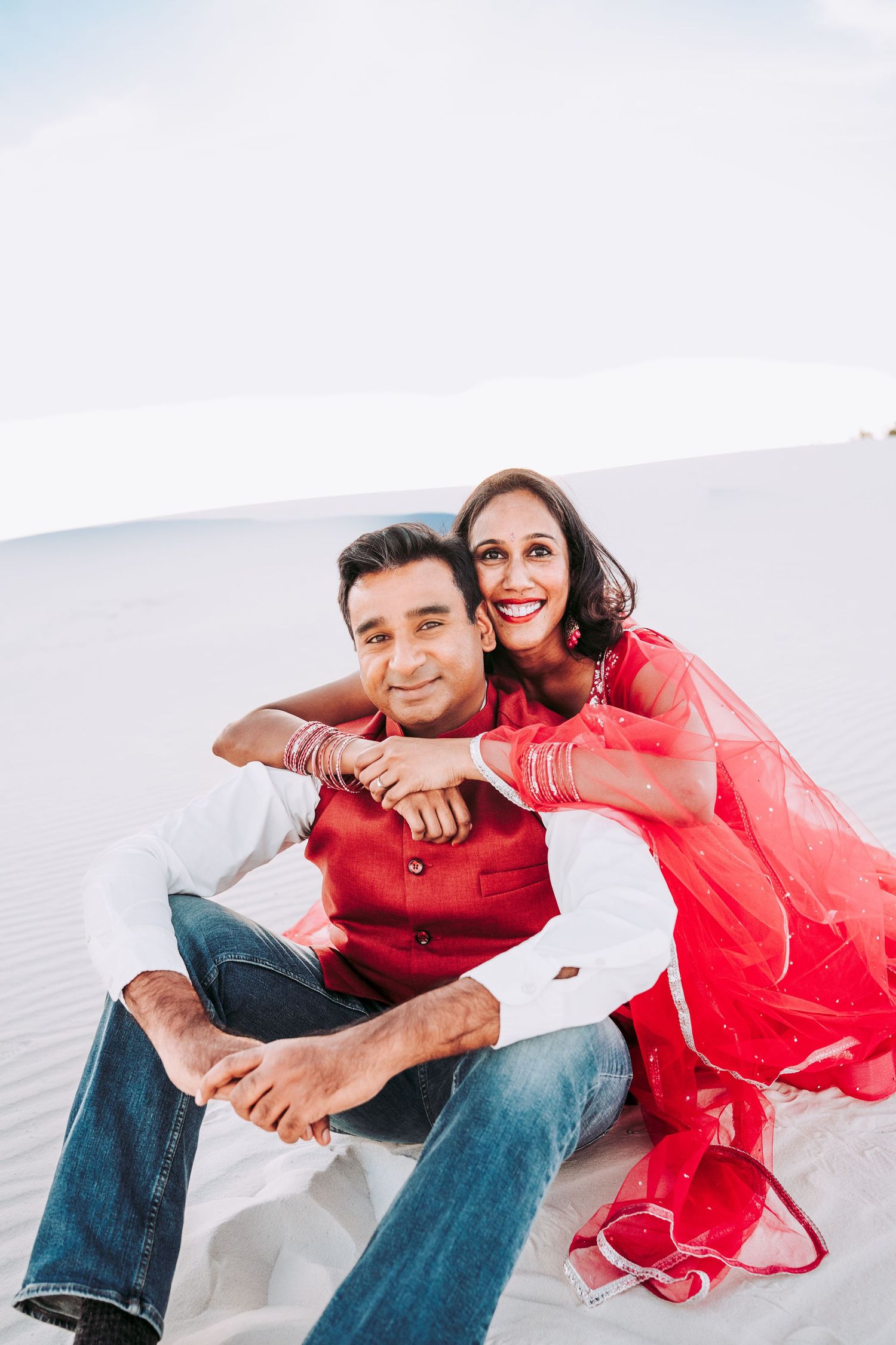 Smiling Indian couple in red and white traditional outfits posing on white sand dunes.