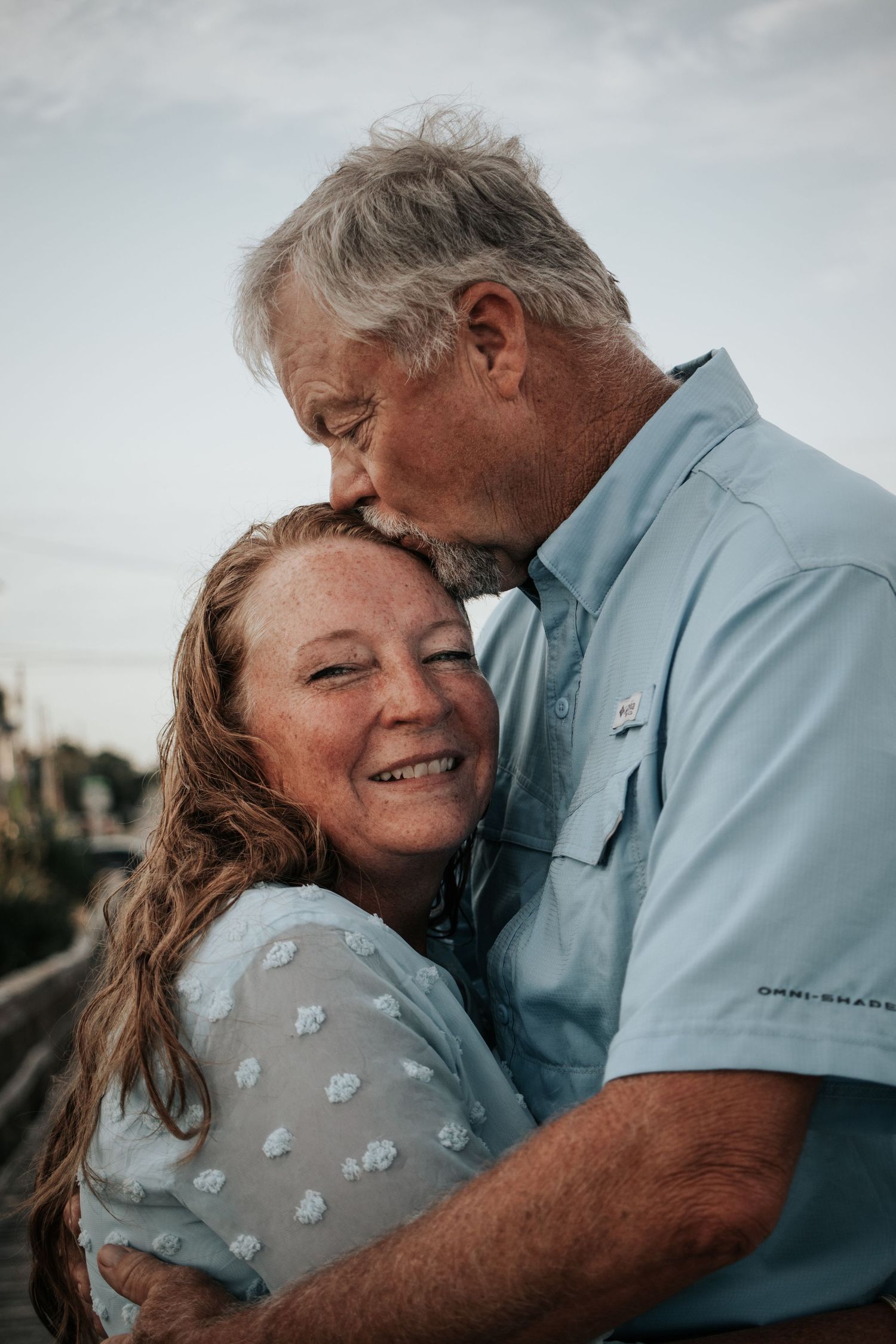 Older couple sharing a tender moment, man kissing woman's forehead on a boardwalk at sunset.