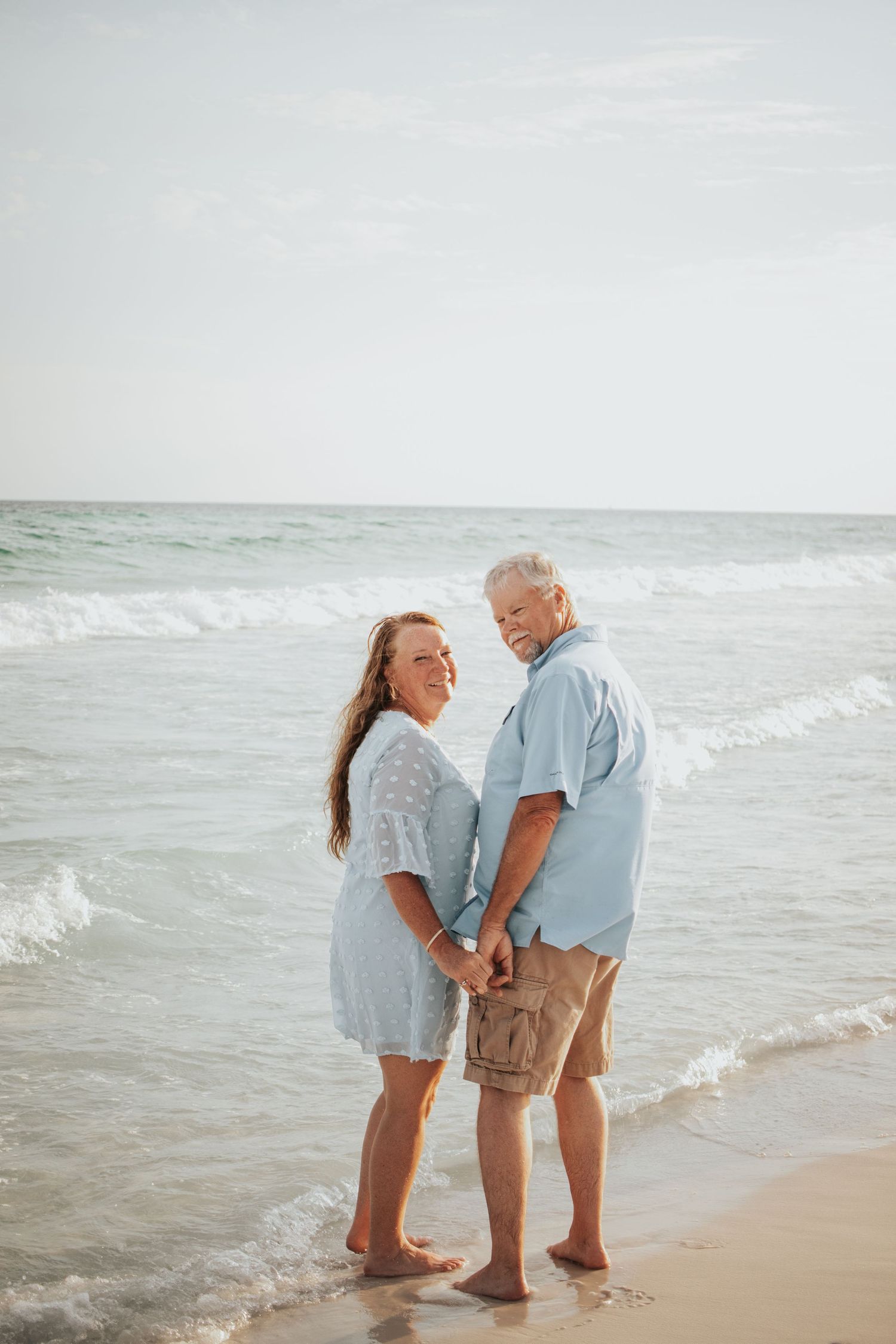 Happy mature couple holding hands and smiling at each other while standing barefoot on a sandy beach.