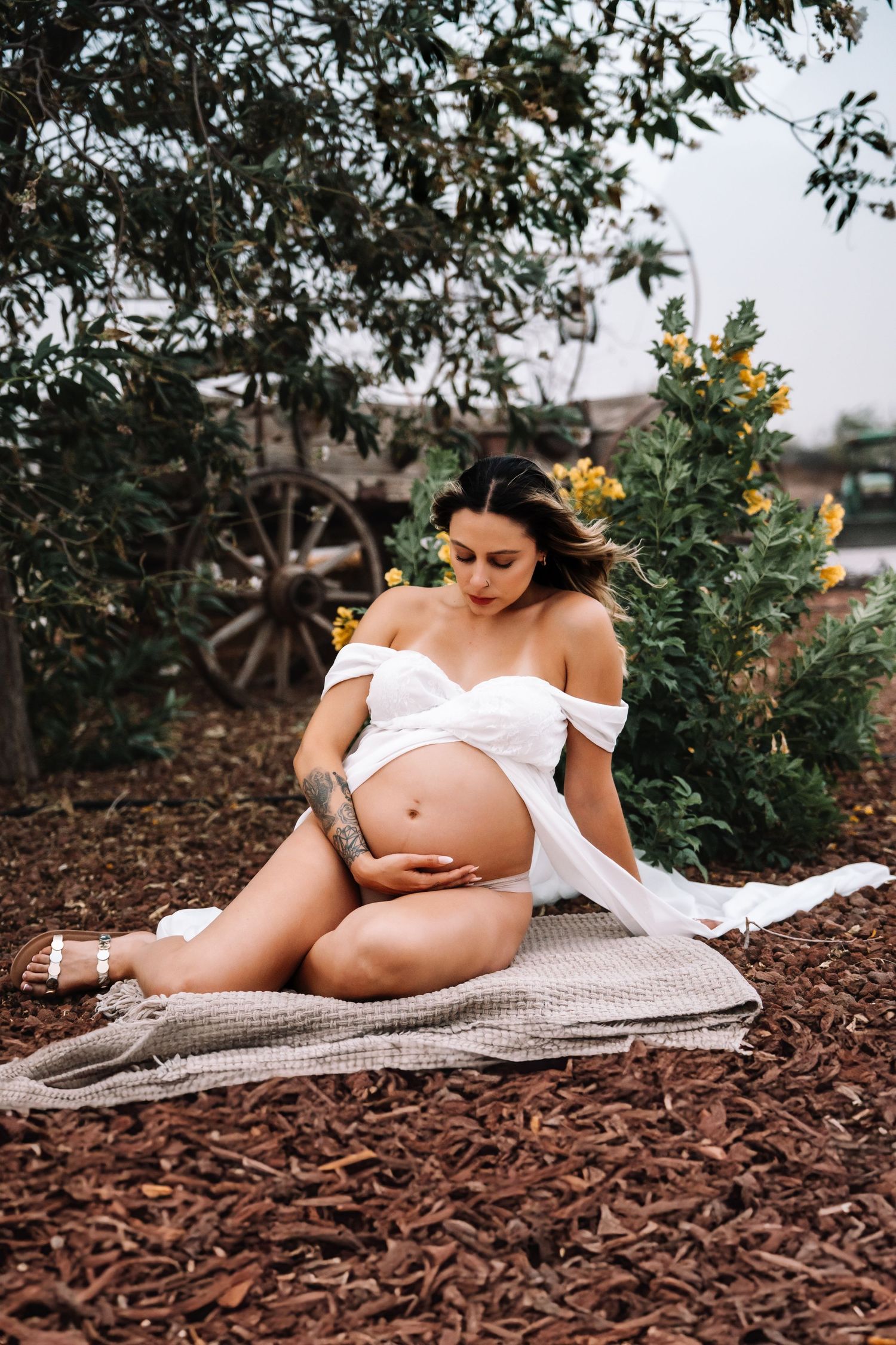 Pregnant woman in white off-shoulder dress sits on blanket outdoors cradling bump near rustic wooden wagon.