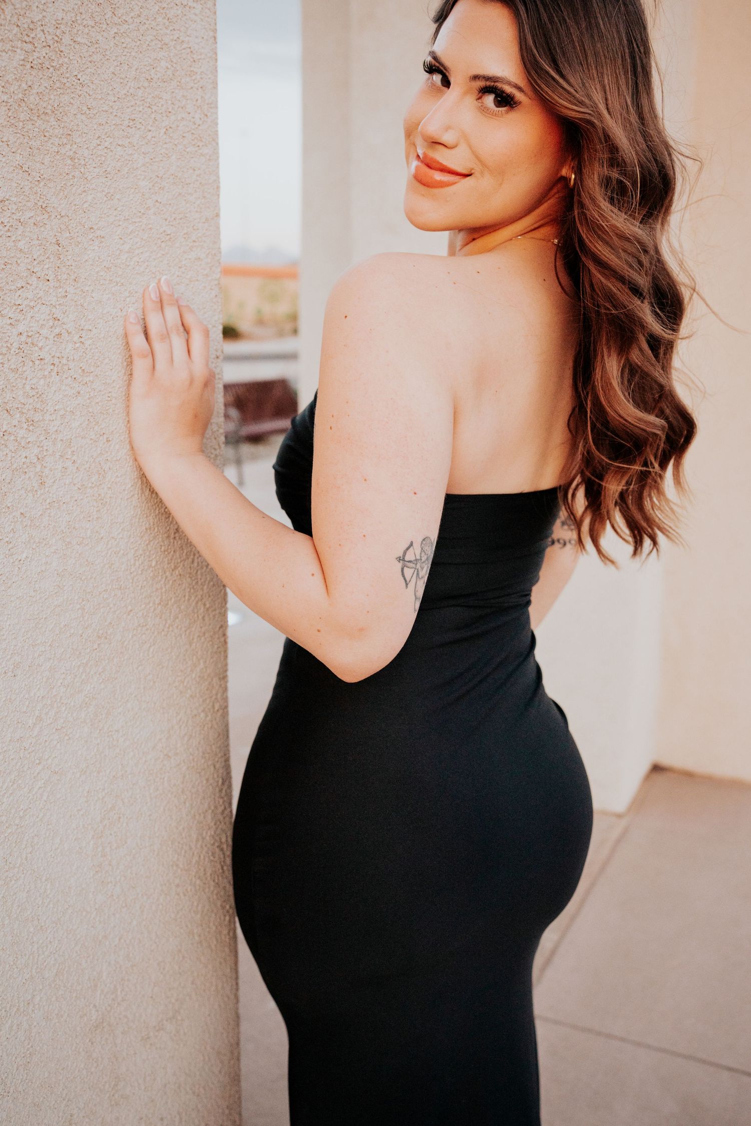 Woman in black strapless dress with tattoo smiling over shoulder against wall.