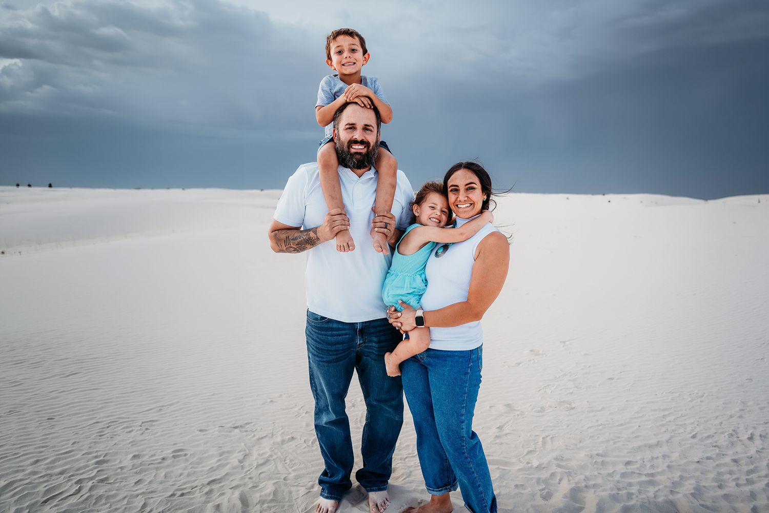 Happy family of four poses together on white sand dunes with stormy skies in the background.