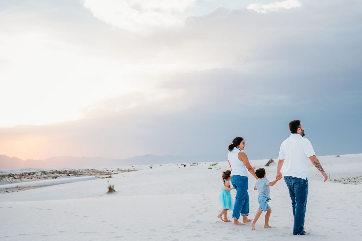 A family of four holds hands while walking across stunning white sand dunes at sunset.