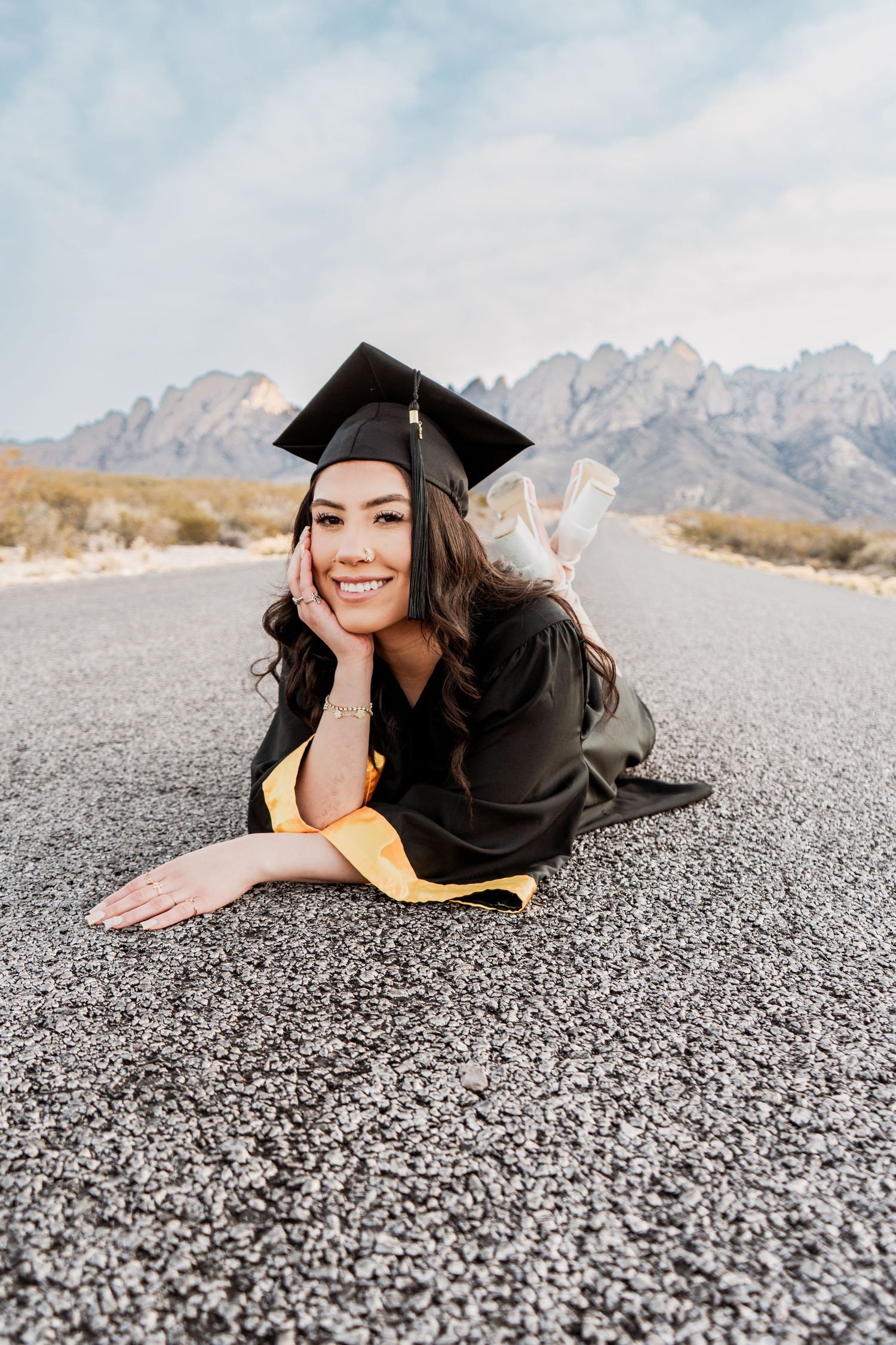 Happy young woman in graduation cap and gown posing on empty road with scenic mountain views.