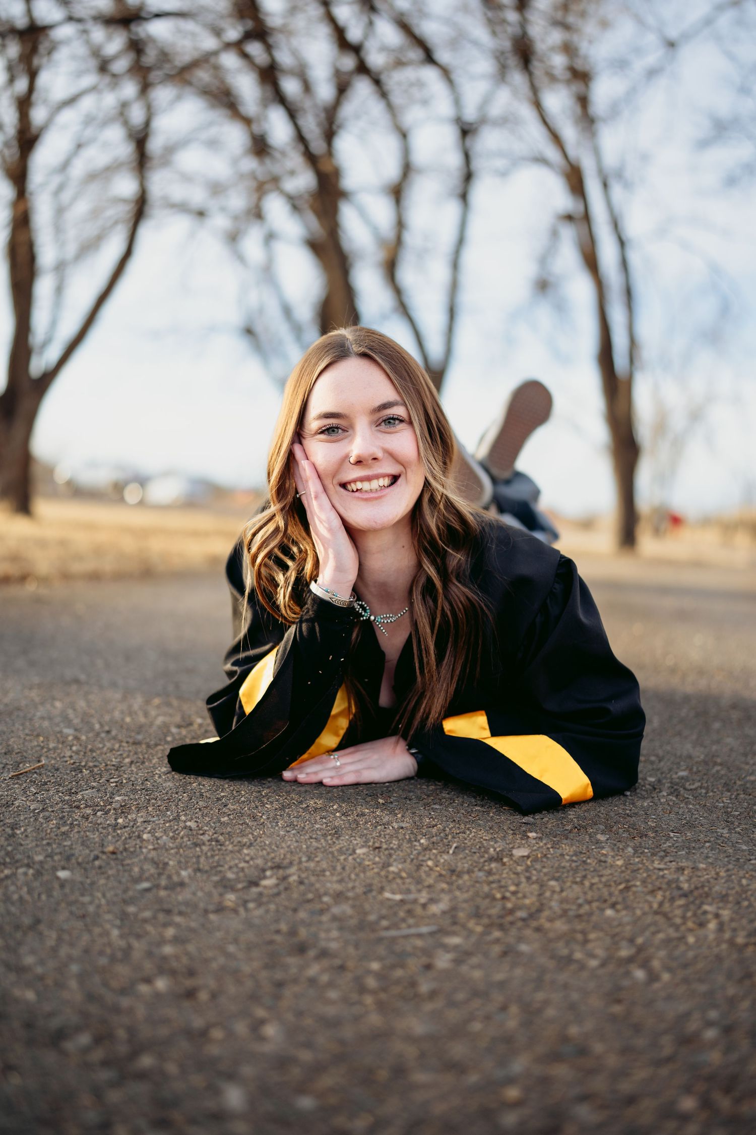 Happy high school senior girl lying on road in black and gold graduation gown smiling at camera.