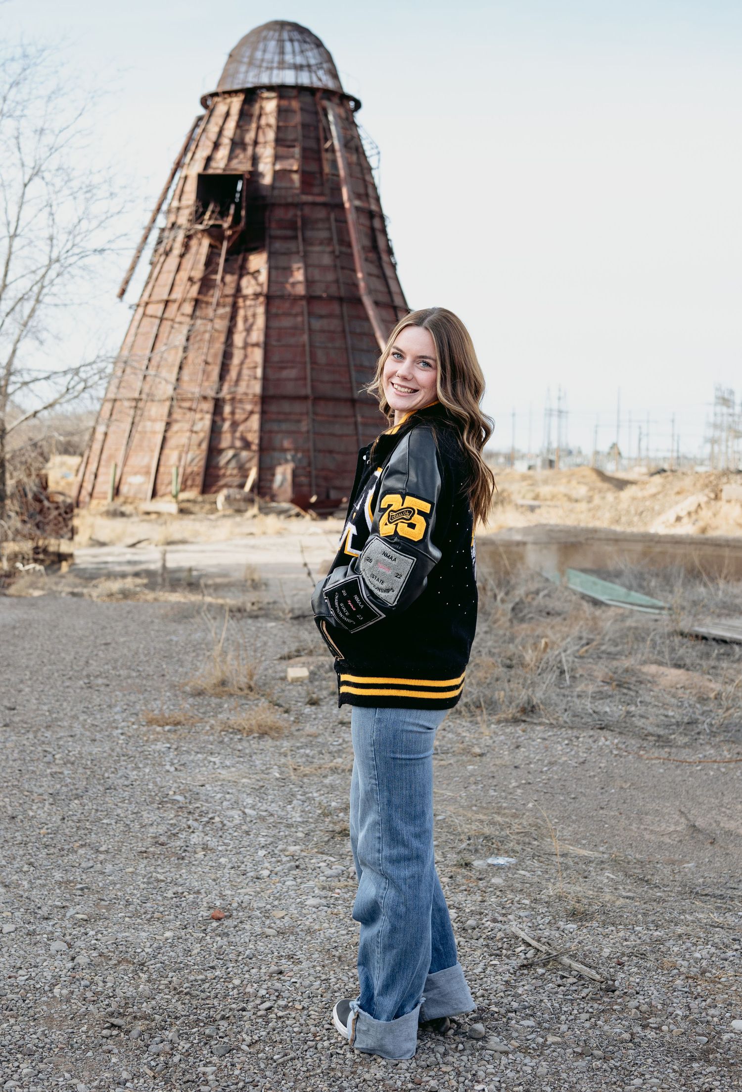 Smiling senior girl in black gold letterman jacket standing before rustic wooden tower structure outdoors.