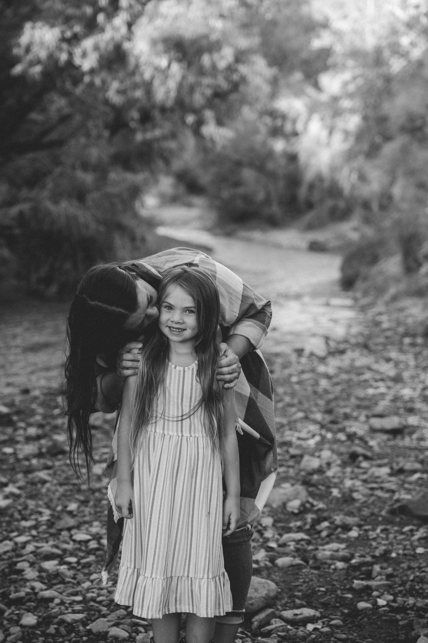 Black and white photo of a woman kissing a young girl on the head outdoors on a rocky path.
