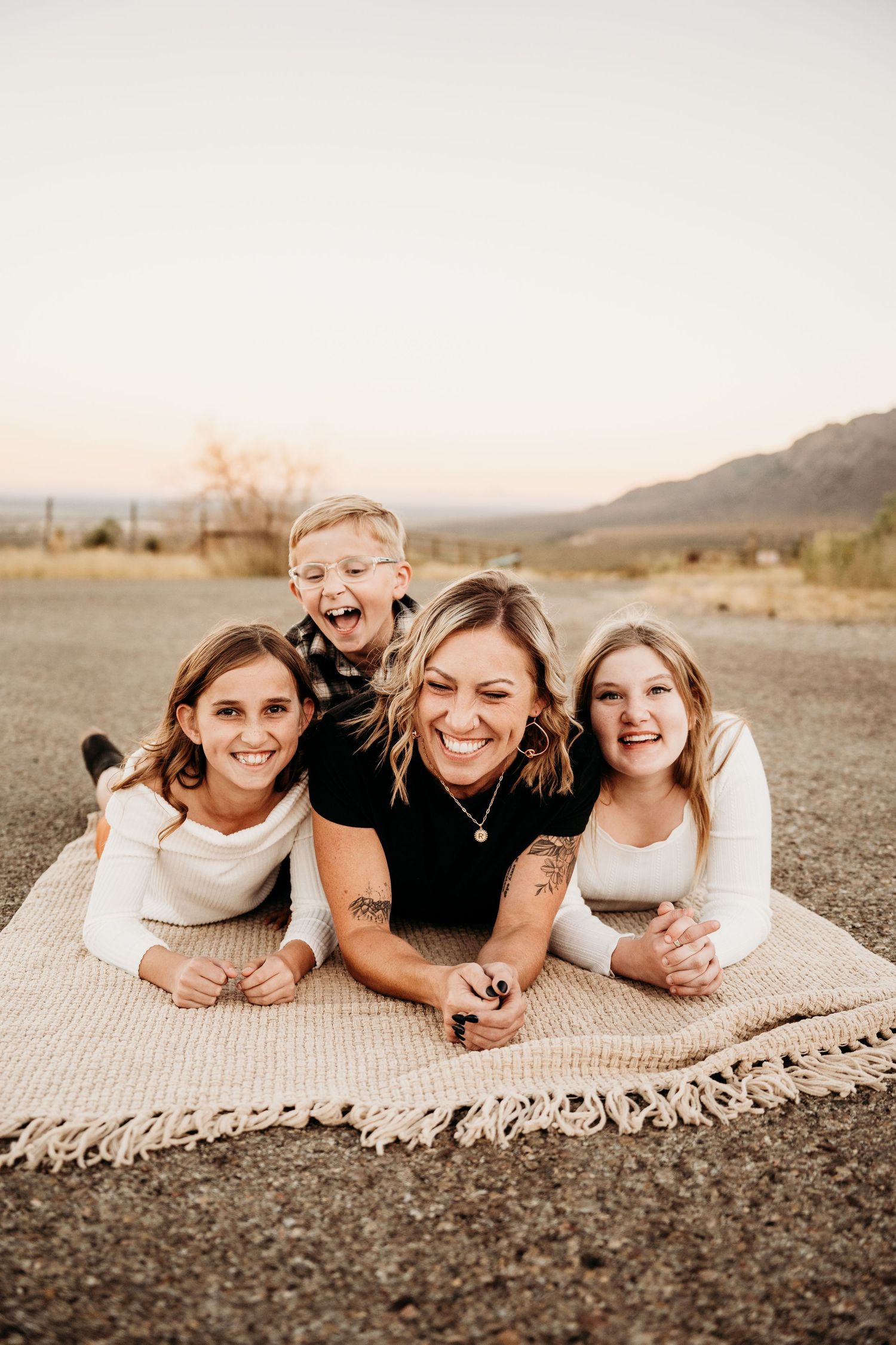 Laughing mother and three children lying on blanket outdoors at sunset.