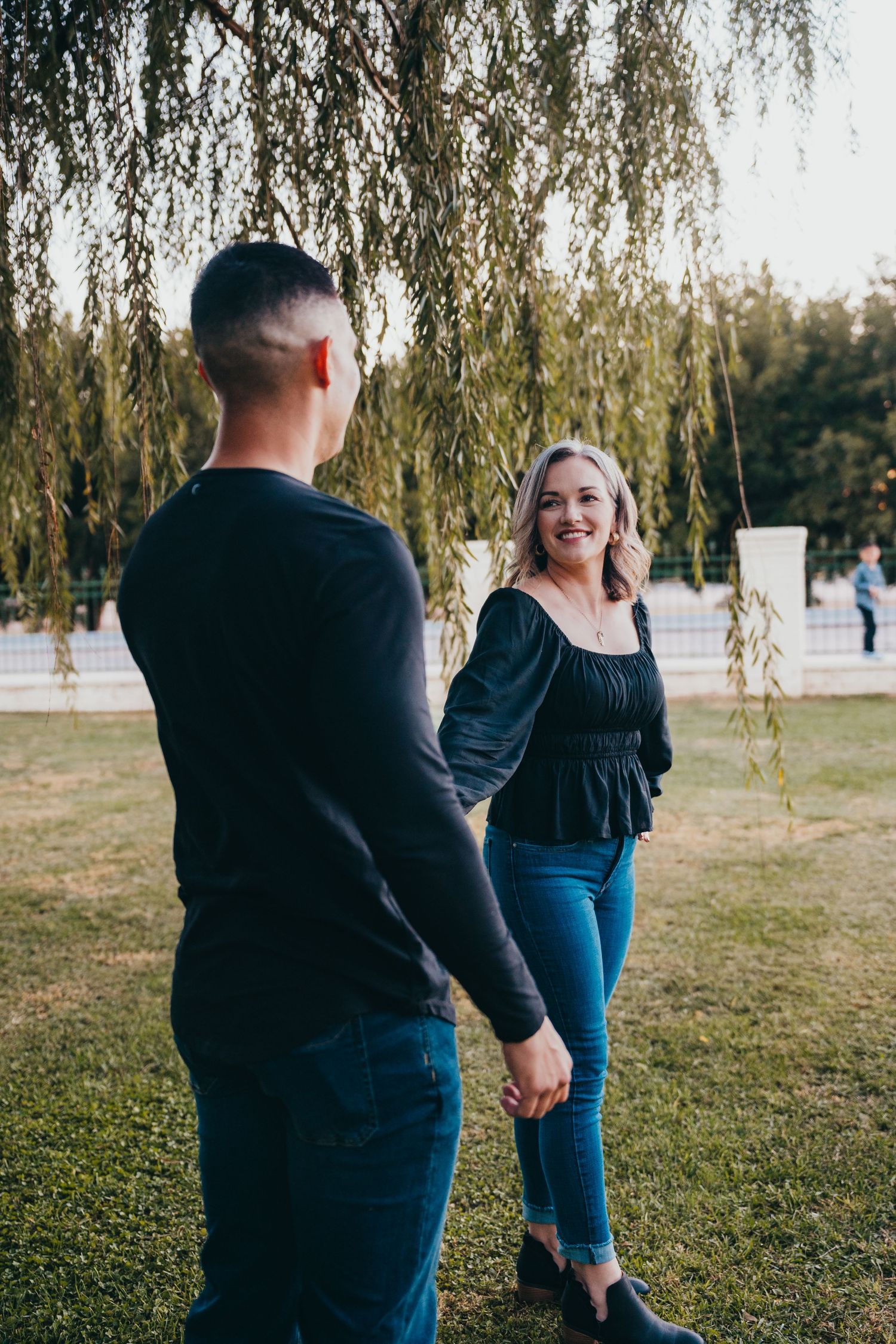 A smiling blonde woman holds hands with a man in a black shirt while standing under a weeping willow tree.