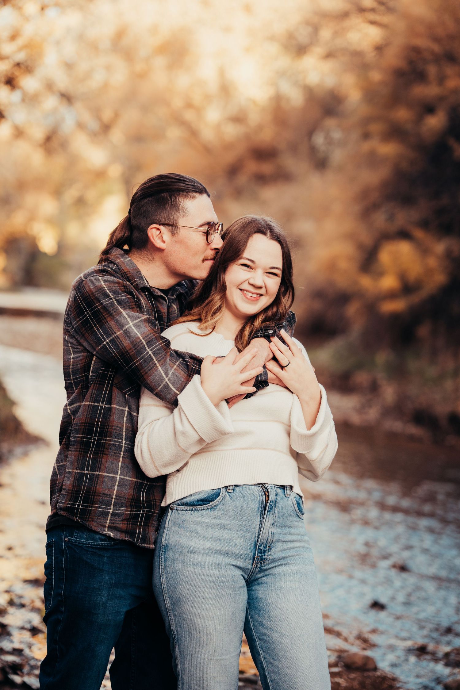 Couple cuddling and laughing together by a rocky stream in fall foliage.