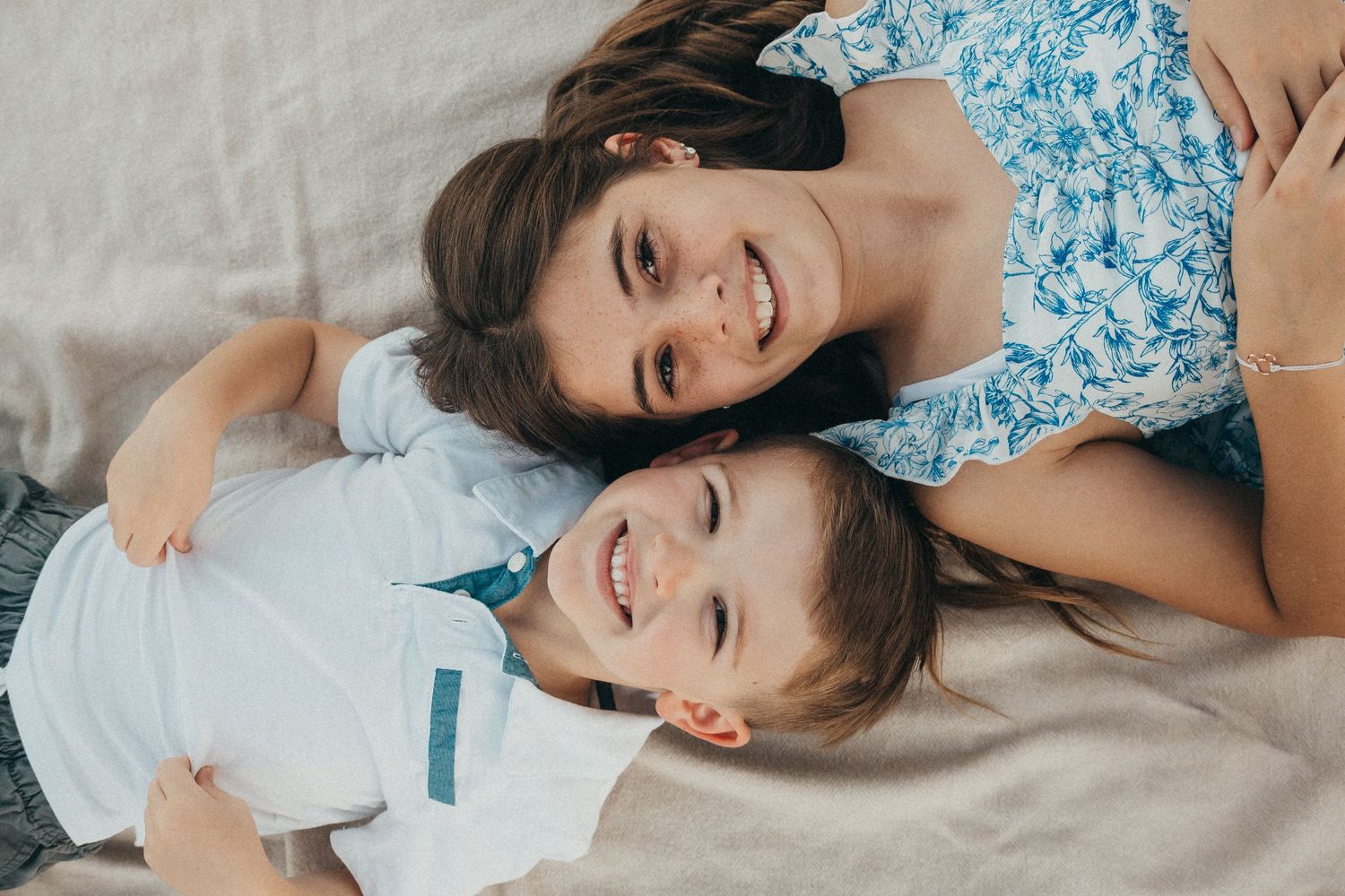 Two siblings lying down together smiling, girl in floral dress hugging younger boy in white shirt.