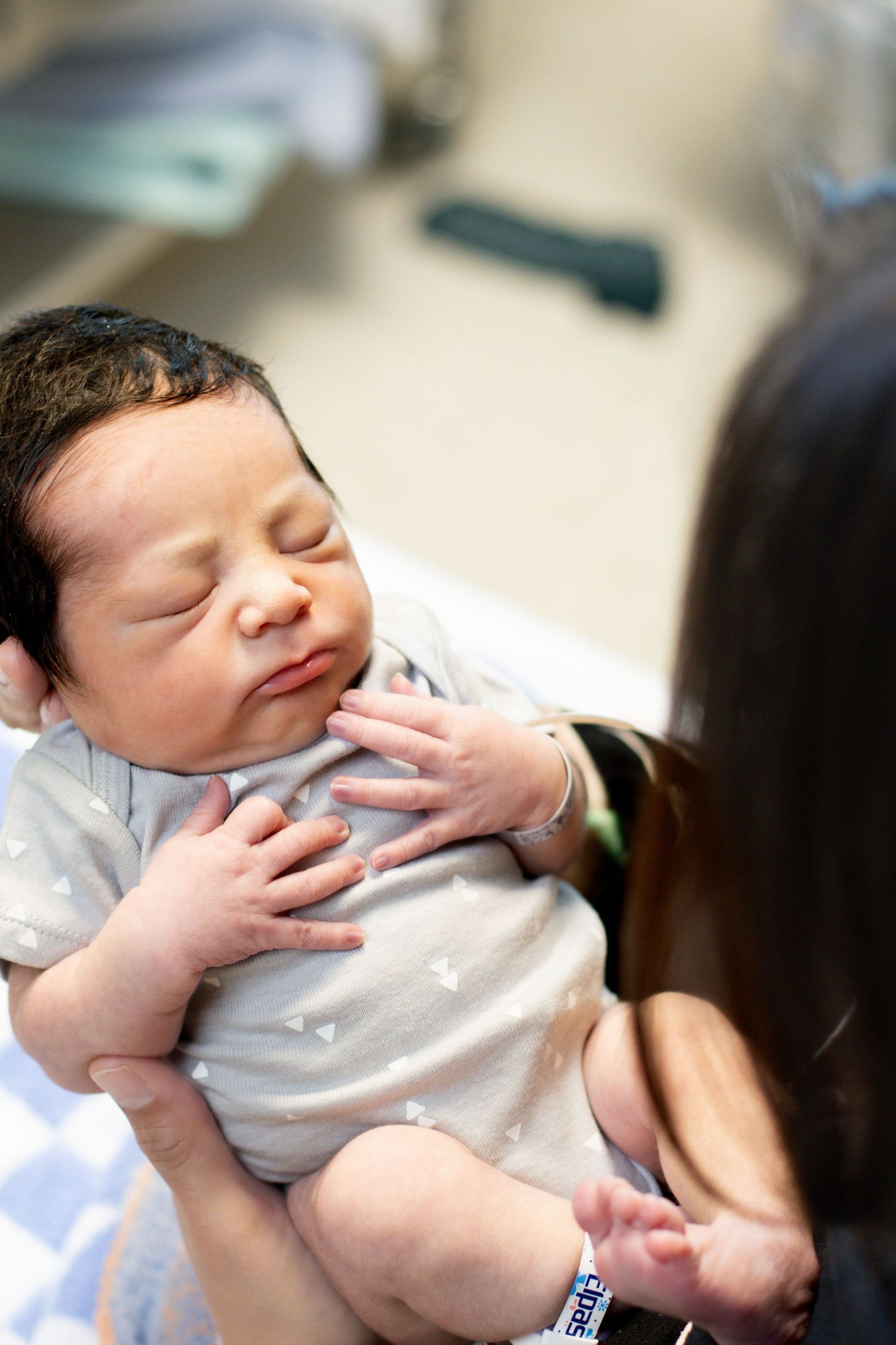 Mother holding sleeping newborn baby with dark hair in hospital room.