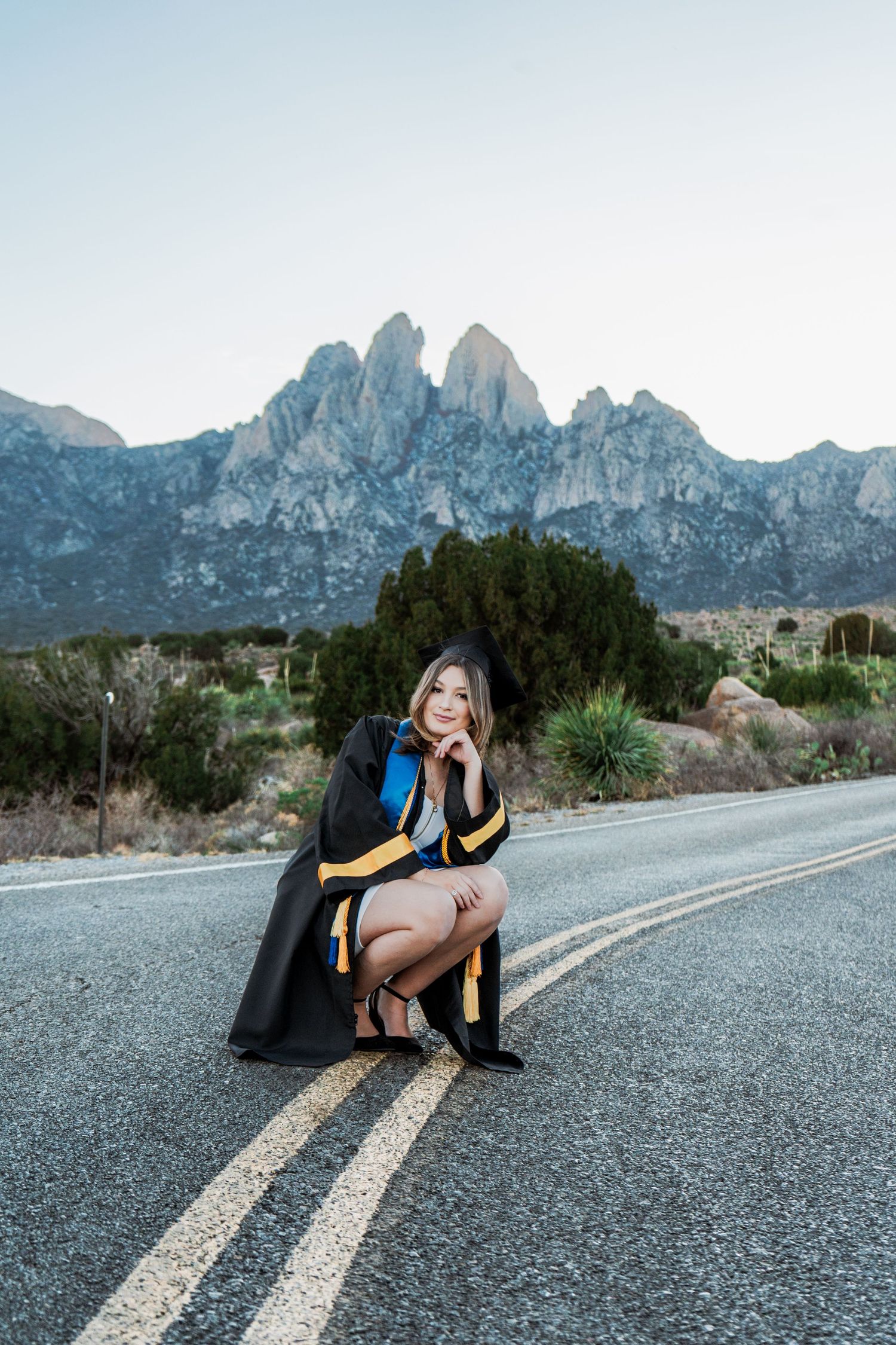 Graduate in black cap and gown posing thoughtfully on road with jagged mountain peaks behind her.
