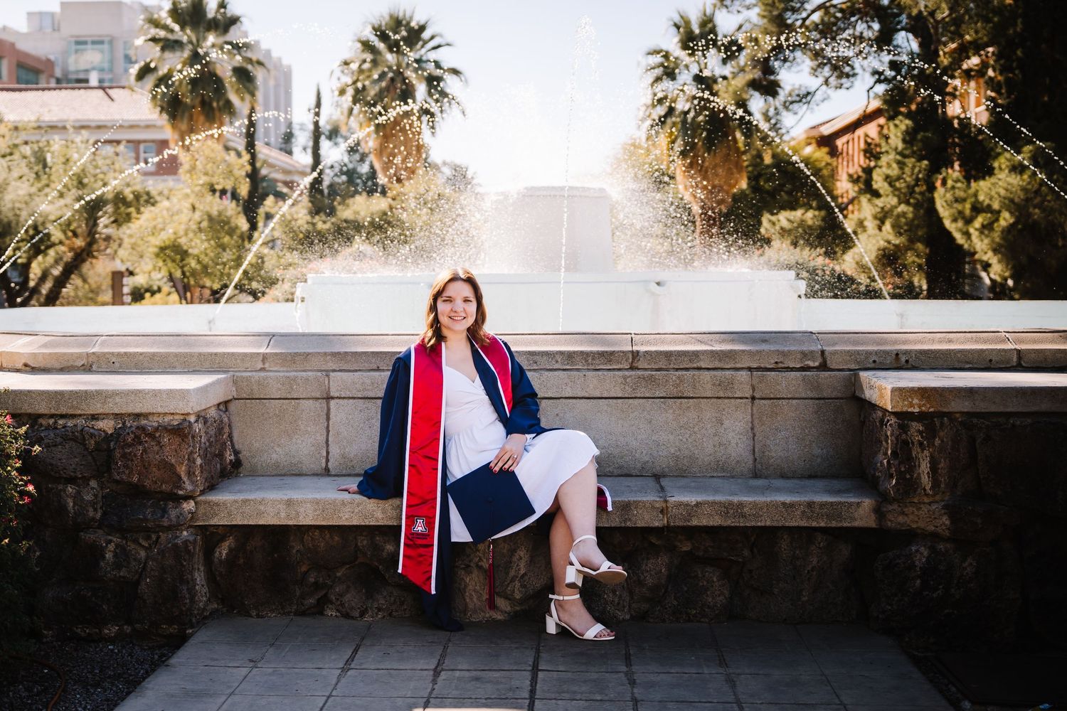 Female graduate in navy cap and gown sitting by a fountain on University of Arizona campus.