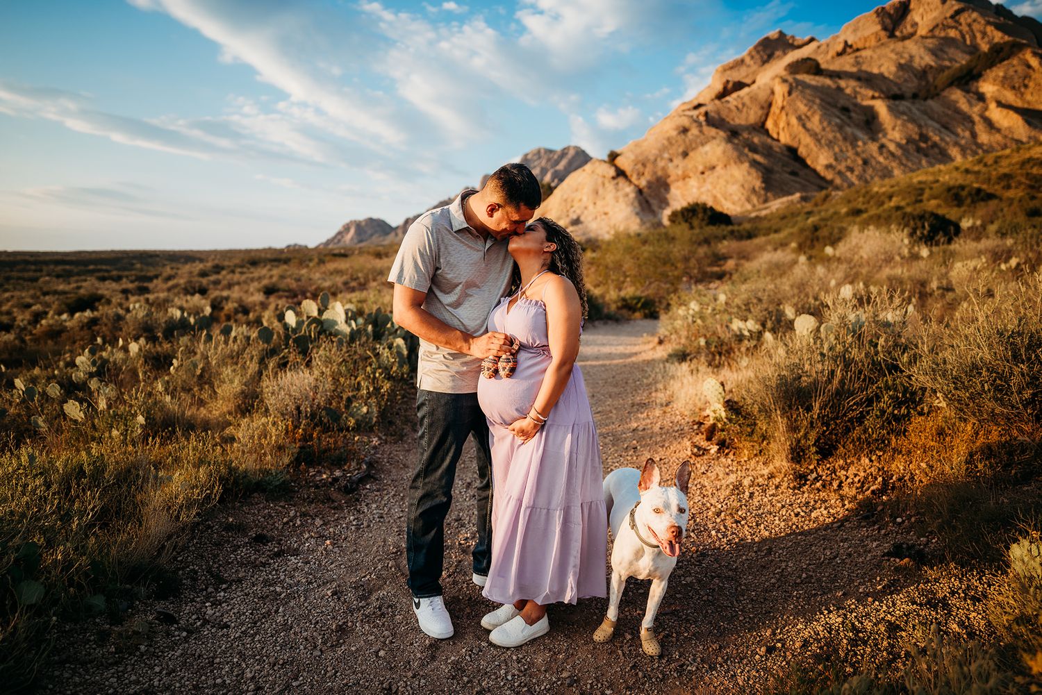 Couple embracing with white dog on a desert trail at golden hour with mountain backdrop.