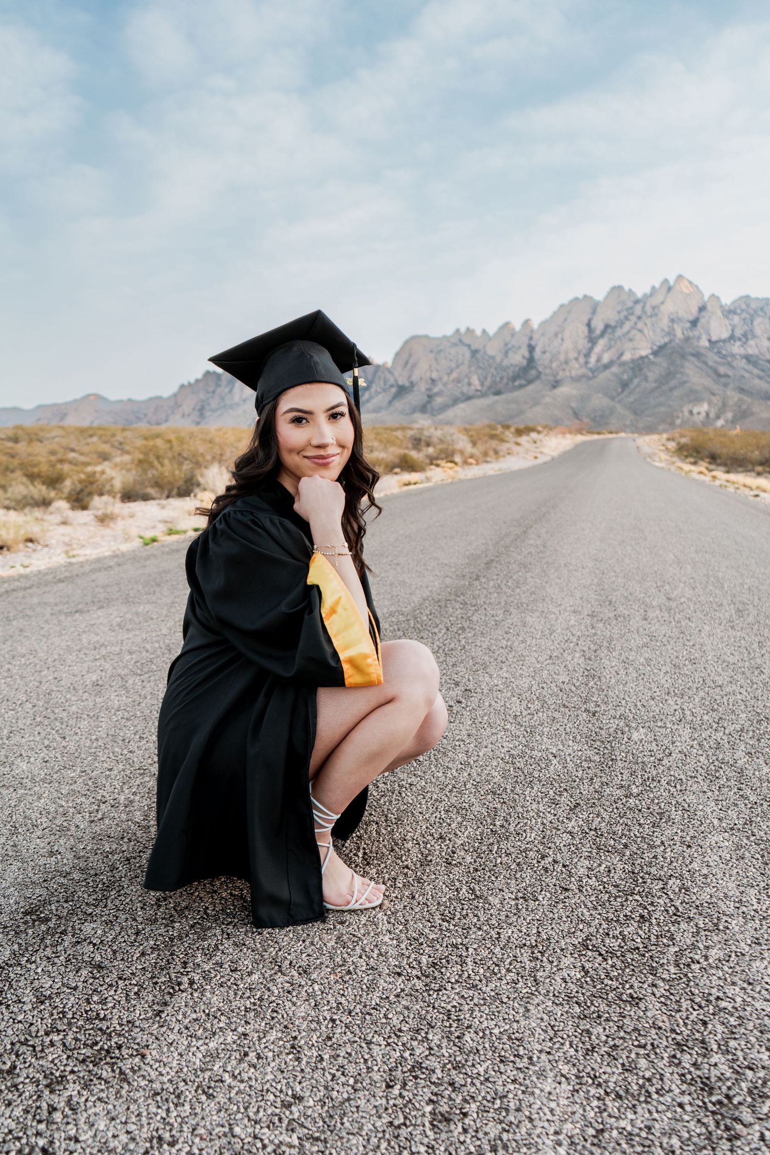 Smiling female graduate in black cap and gold gown crouches on a desert road with mountain scenery behind her.