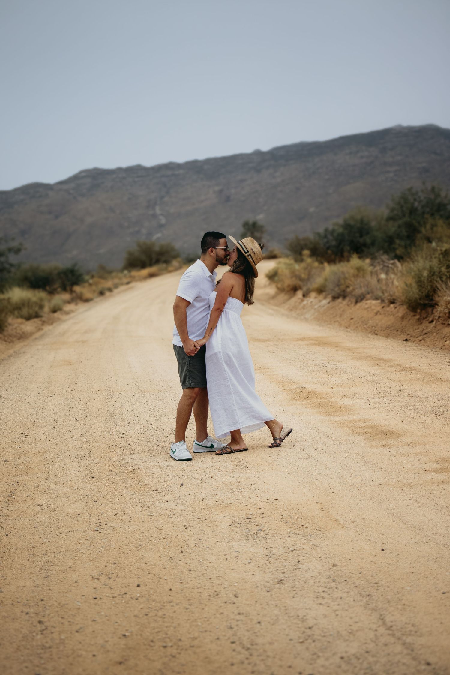 Couple kissing on a sandy desert road with mountain backdrop, woman wearing straw hat and white dress.