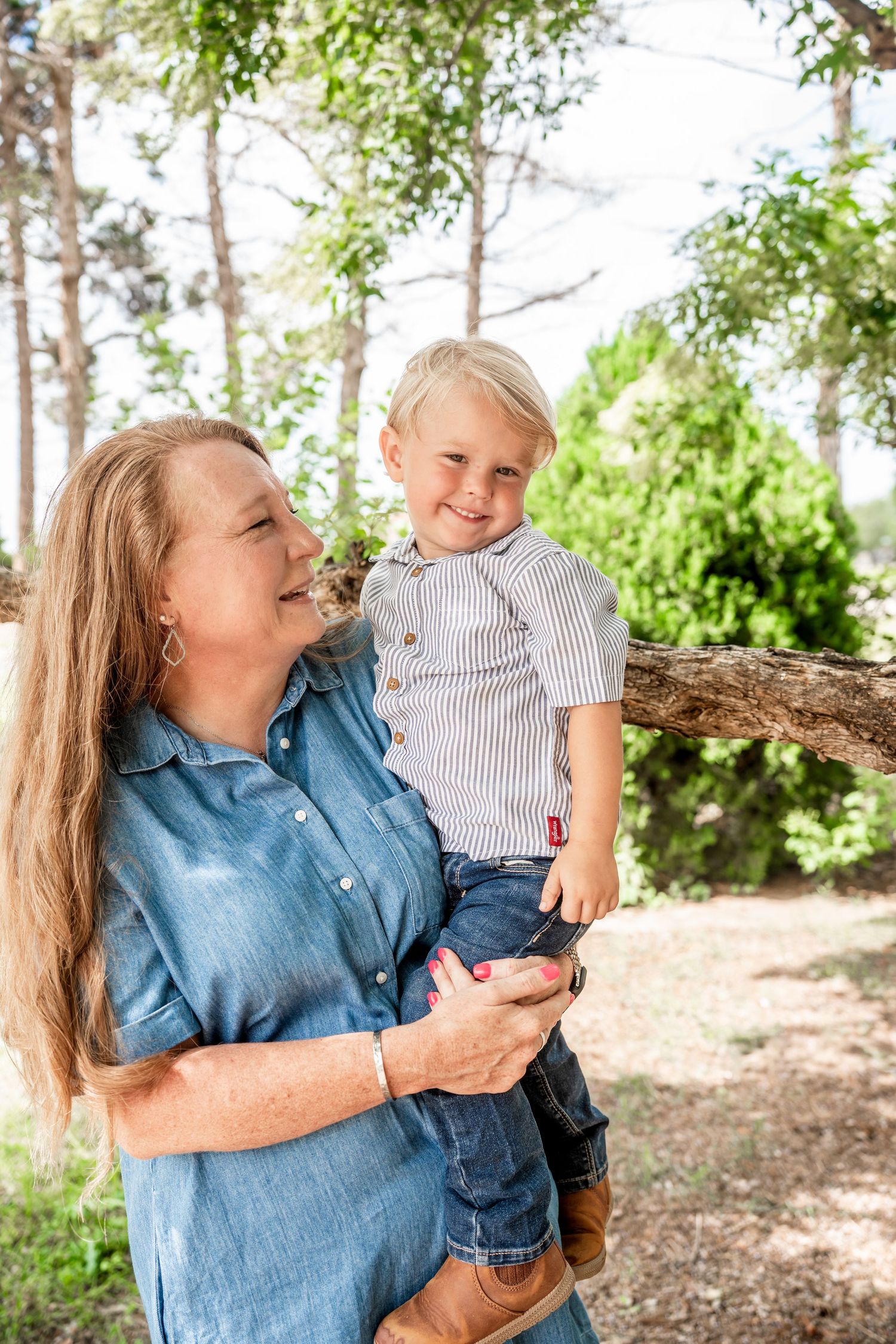 Grandmother in blue dress holding laughing toddler boy outdoors in wooded park setting.