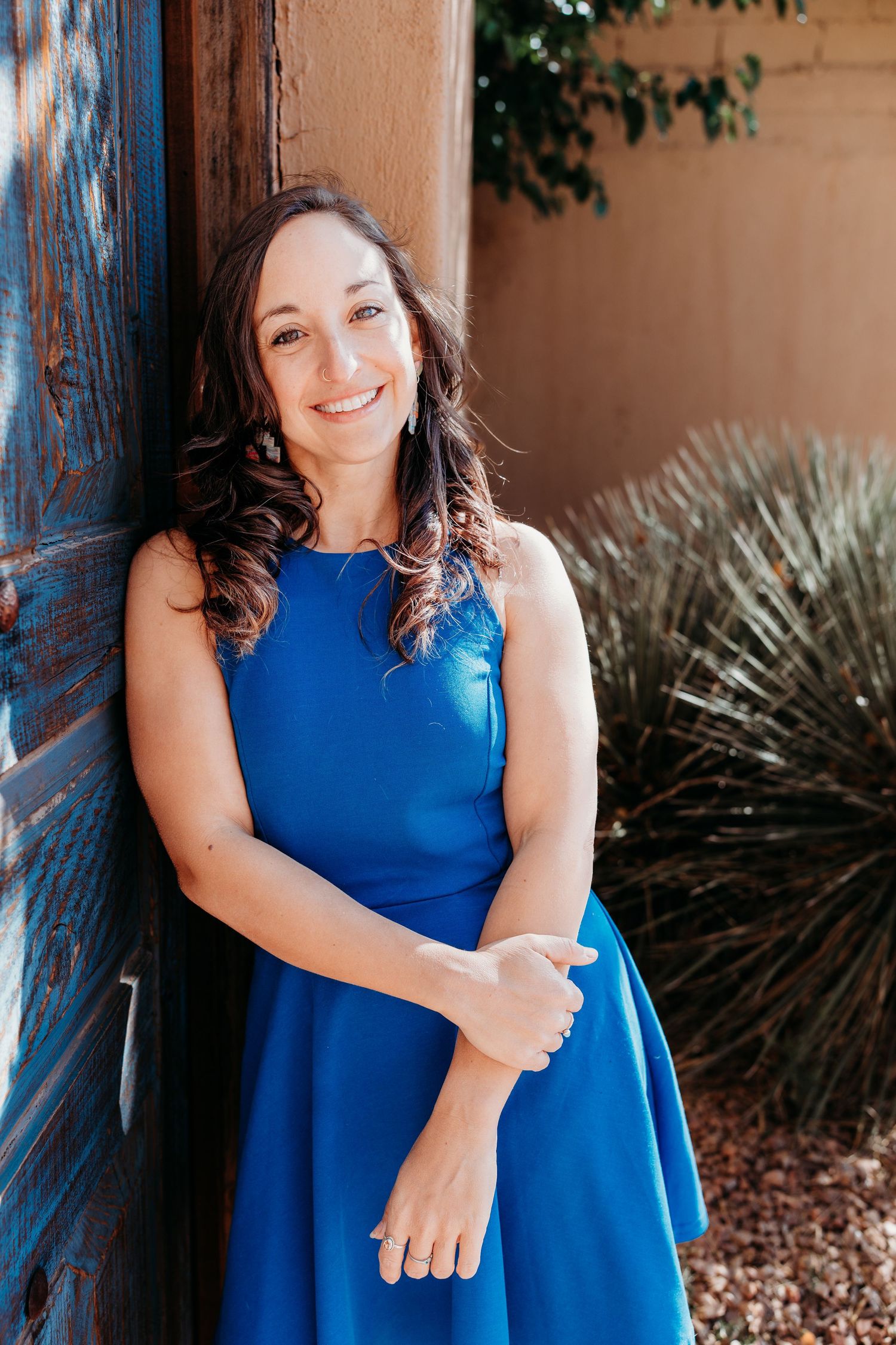 Brunette woman in blue dress smiling while leaning against rustic blue wooden door.
