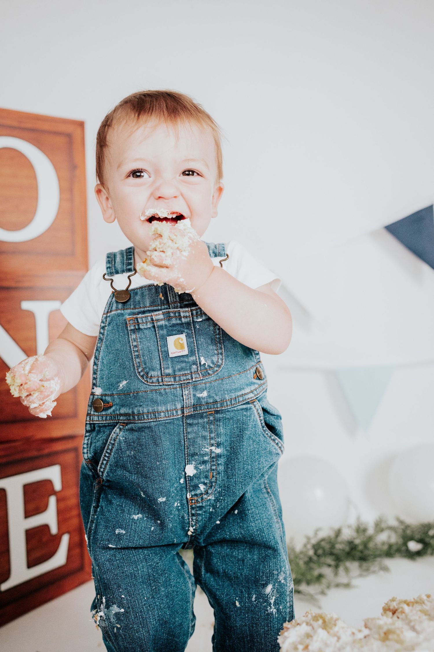 Happy toddler boy in Carhartt denim overalls laughing while eating birthday cake at a cake smash photo session.