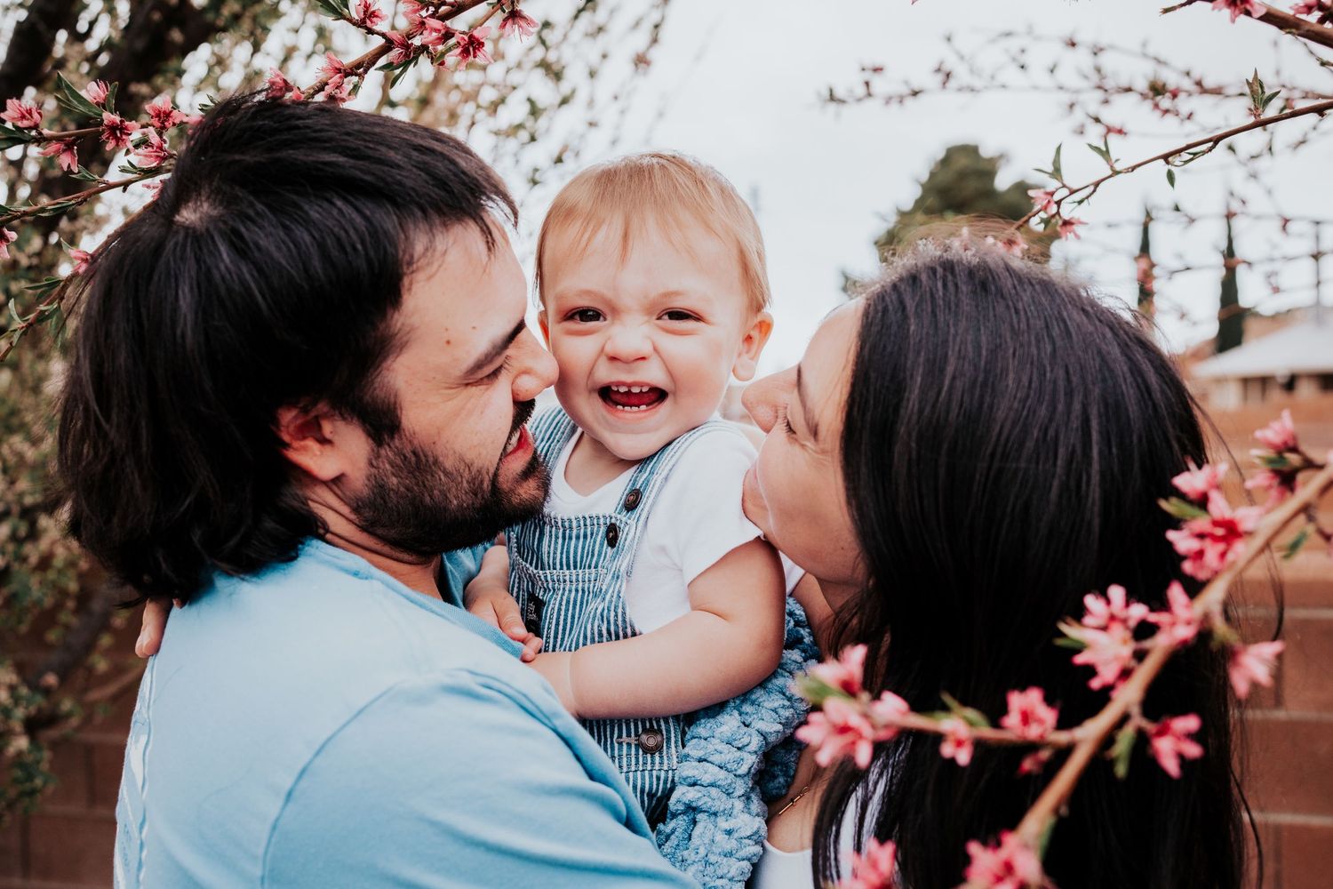Two parents kissing their giggling baby boy on each cheek near blooming pink spring blossoms.