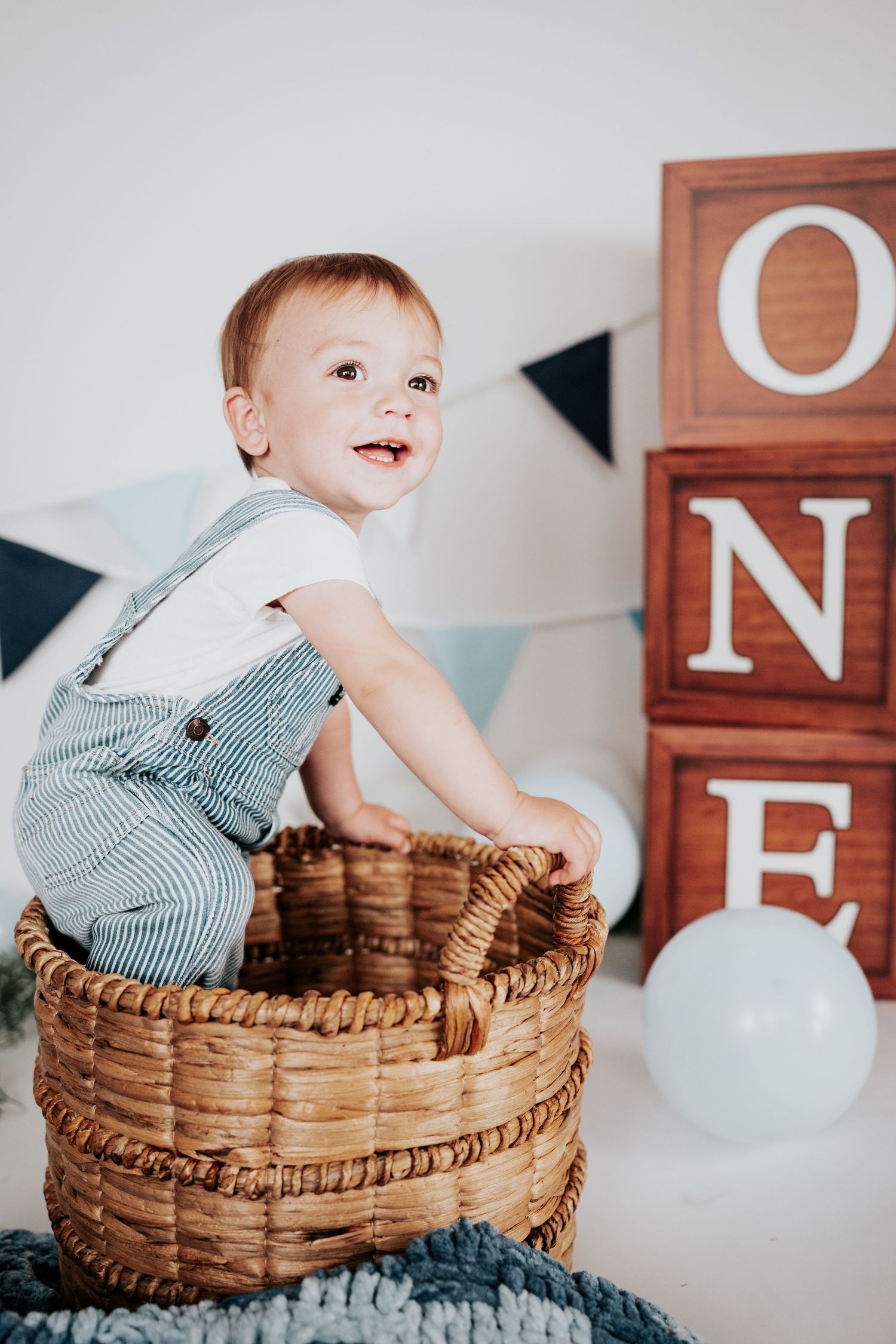 Smiling one-year-old boy in striped overalls posing in wicker basket beside wooden ONE birthday blocks backdrop.