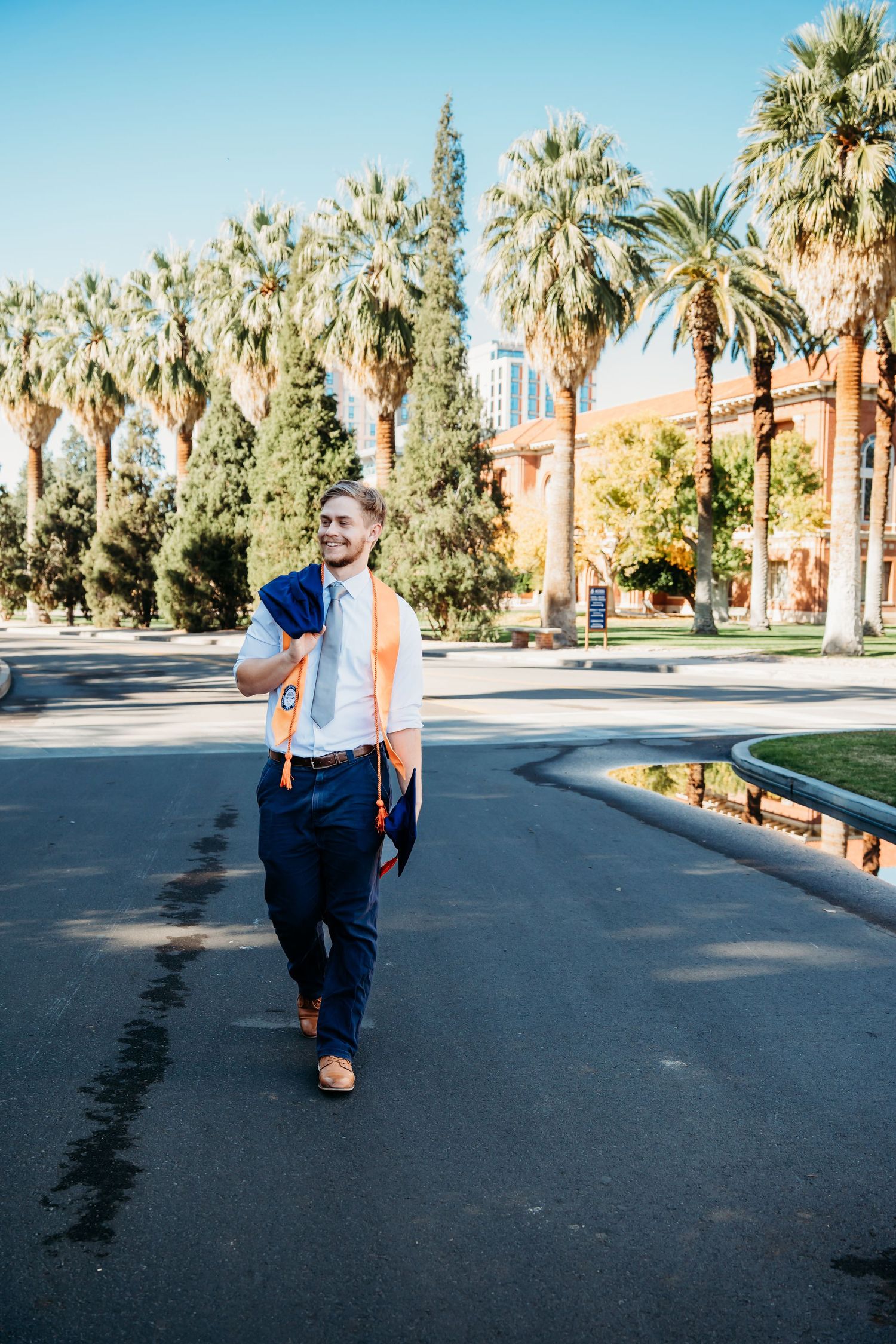 Male graduate in blue suit with orange sash walking on palm tree-lined university campus.