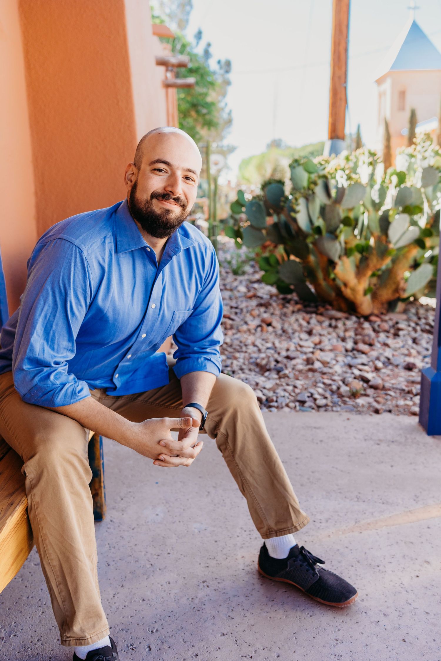 Bald man with beard in blue shirt sitting outdoors near desert cactus garden.