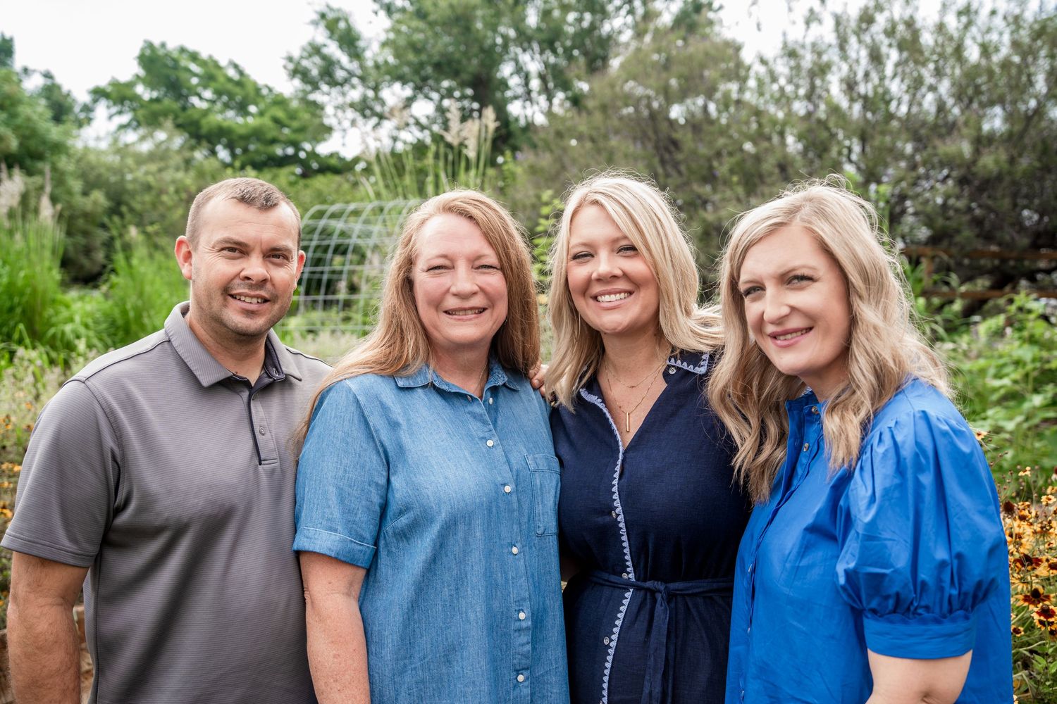 Four adults including two women and two men smile together in a bright outdoor garden setting.