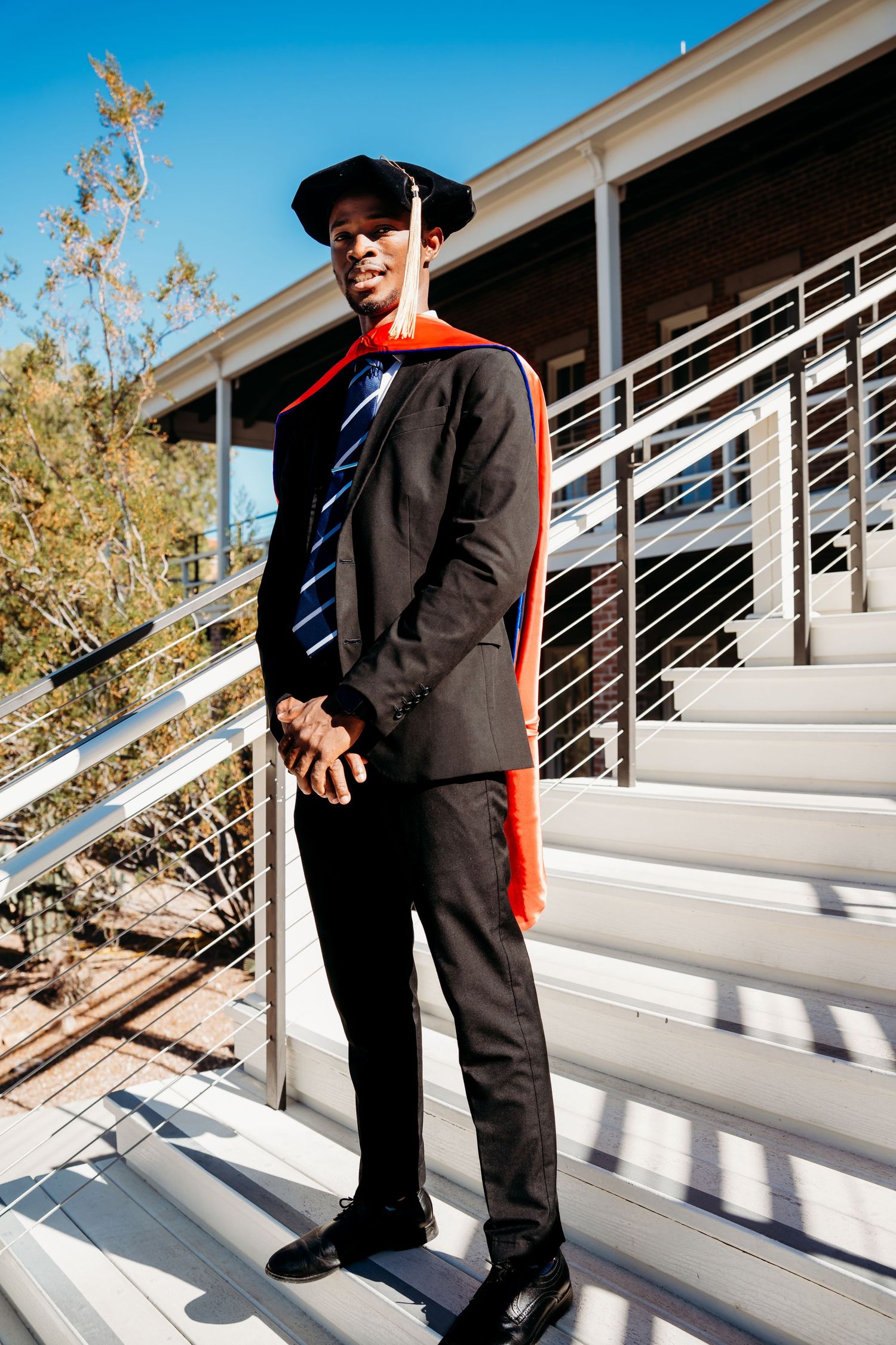 A young man in a dark suit, tie, and graduation cap with red academic hood stands on outdoor stairs under a blue sky.