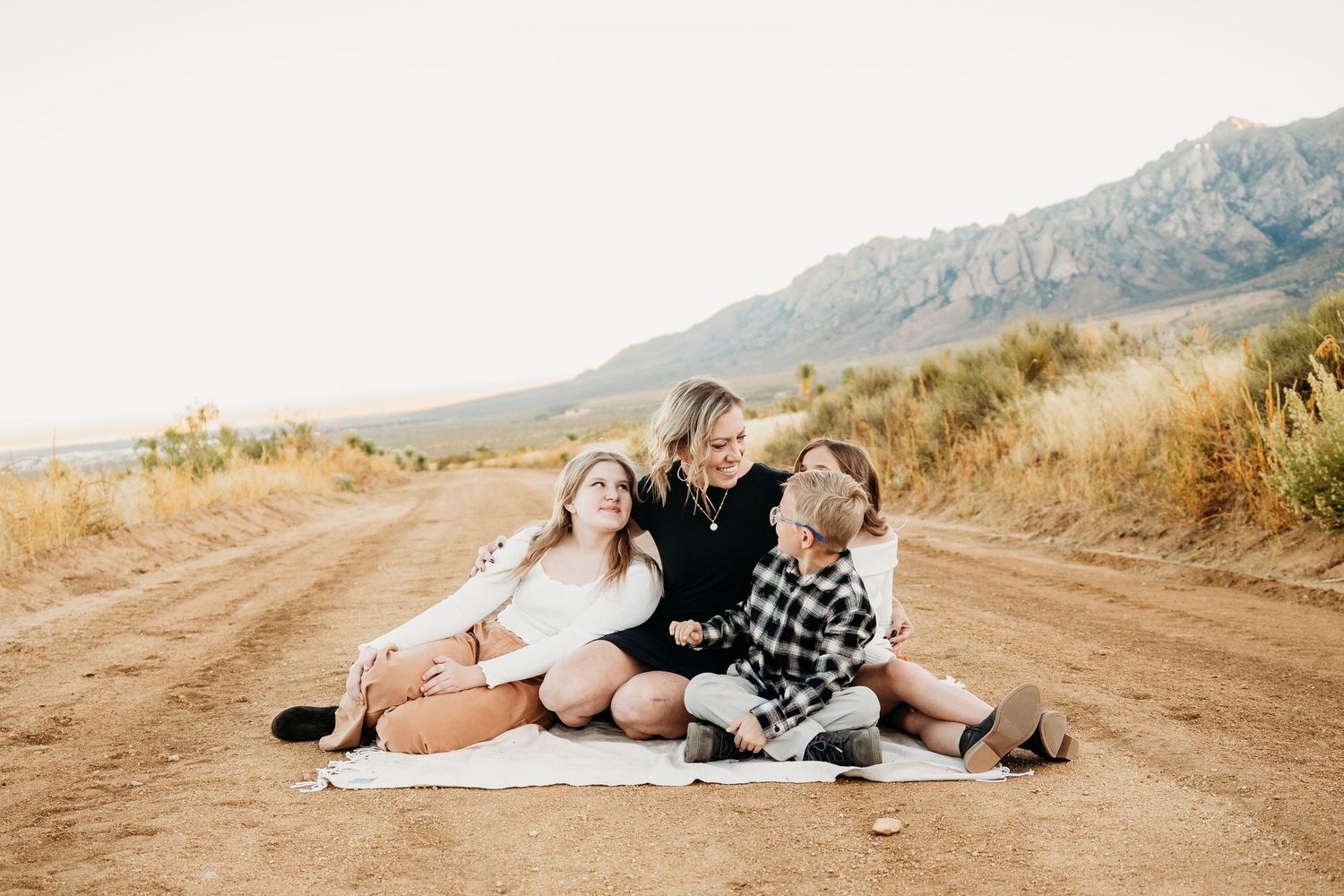 A mother and her two children sit together on a blanket on a sunny desert road.