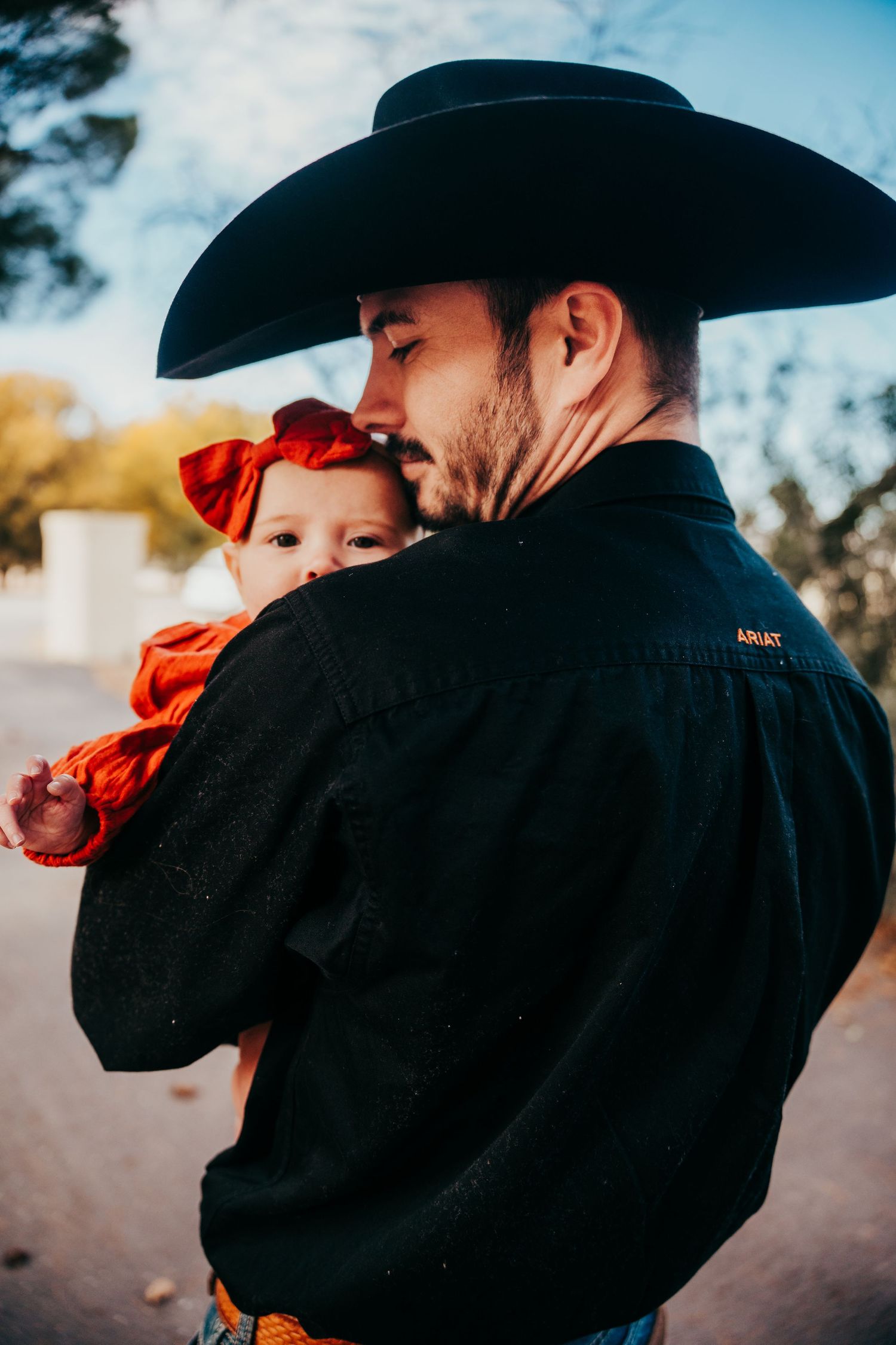 Father in black cowboy hat and Ariat shirt tenderly holding baby girl in red outfit and bow.