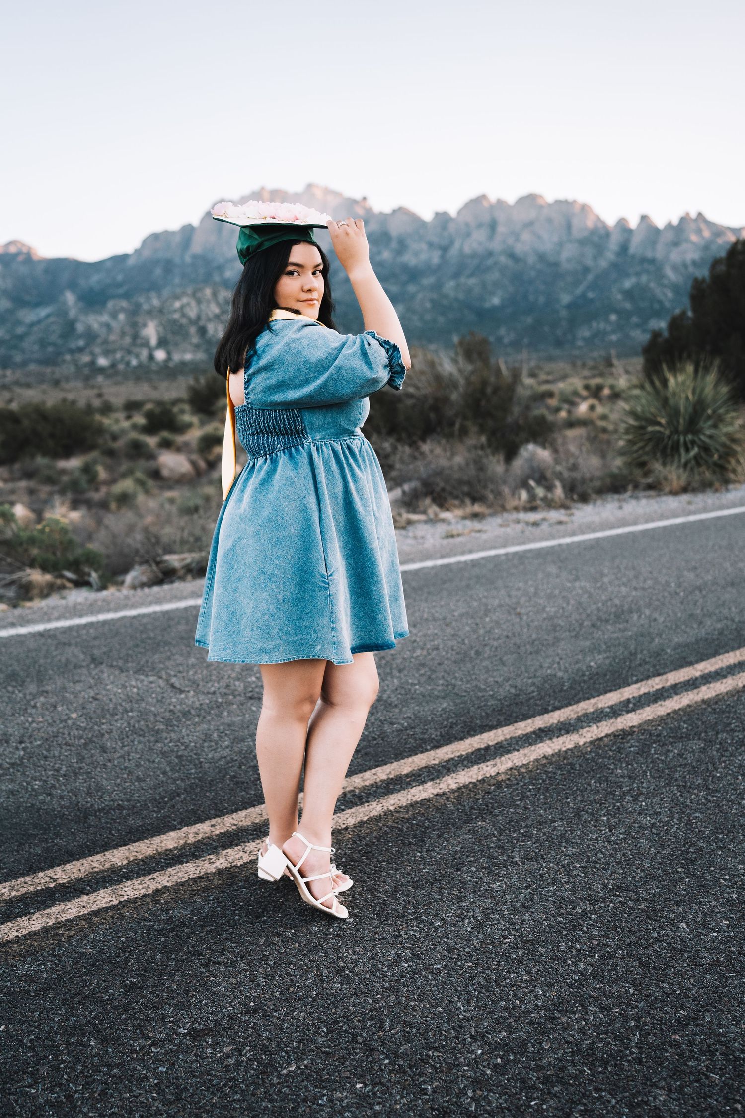 Young female graduate in a denim dress balancing her decorated graduation cap on a scenic desert road.