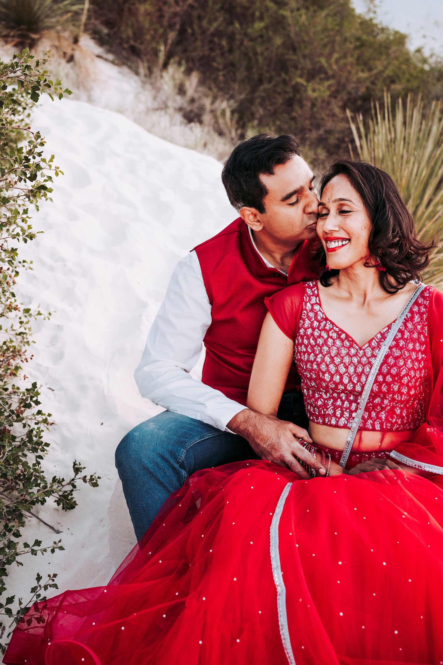Indian couple in red traditional attire sharing a tender kiss on white sand dunes.