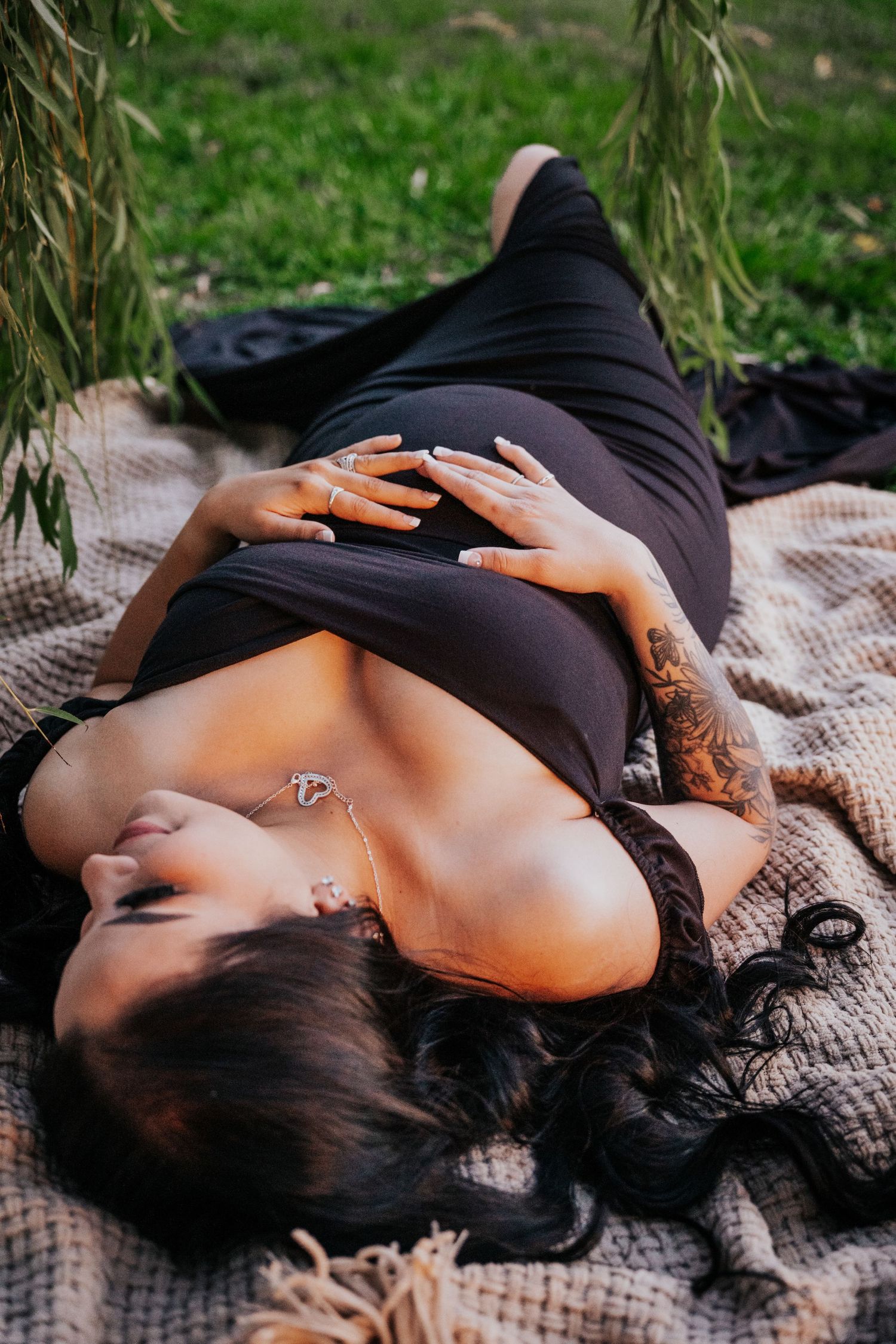 Tattooed pregnant woman in black dress lying on blanket cradling bump under willows.