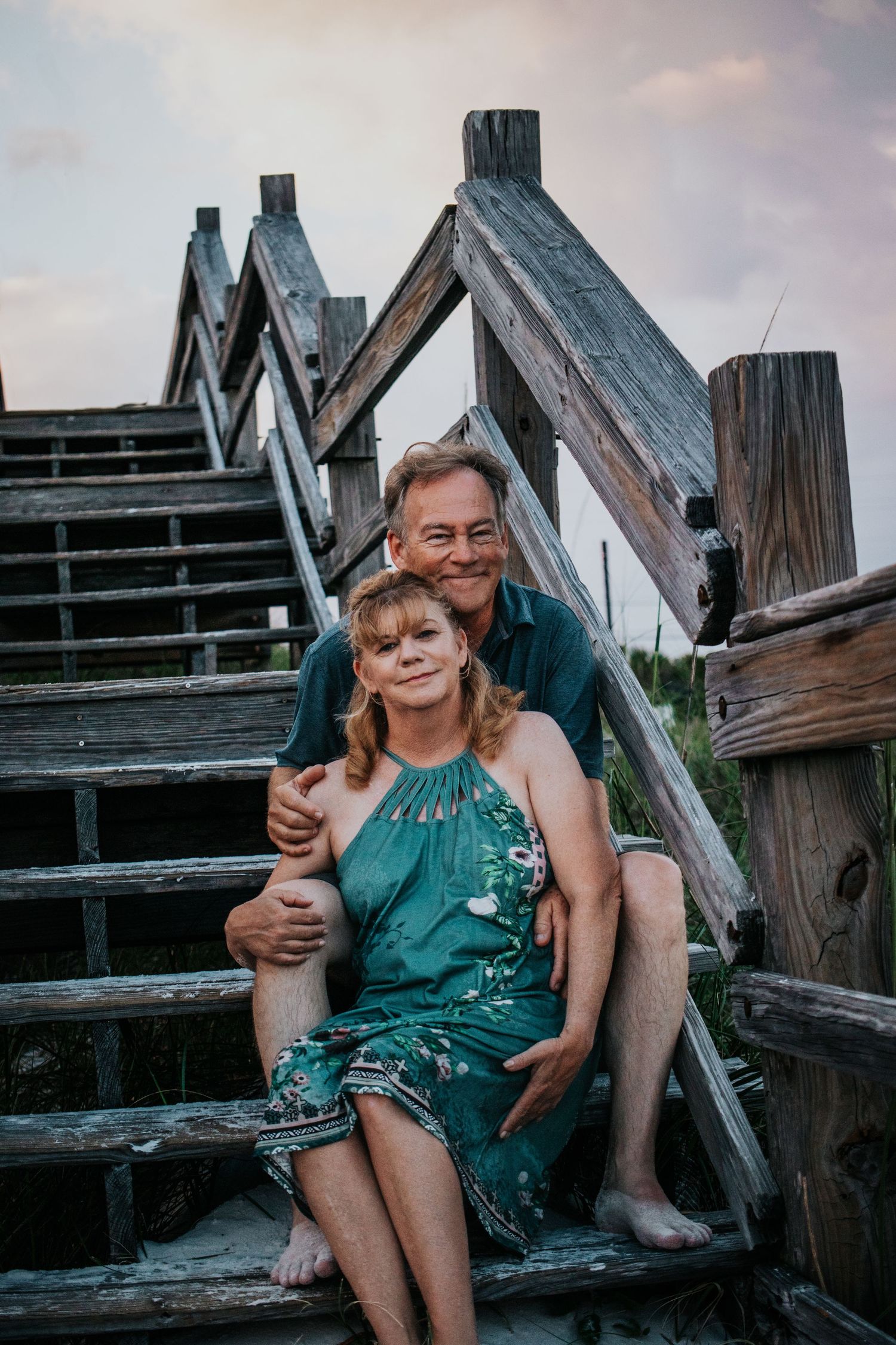 Couple sitting together on weathered wooden beach stairs smiling at the camera at dusk.