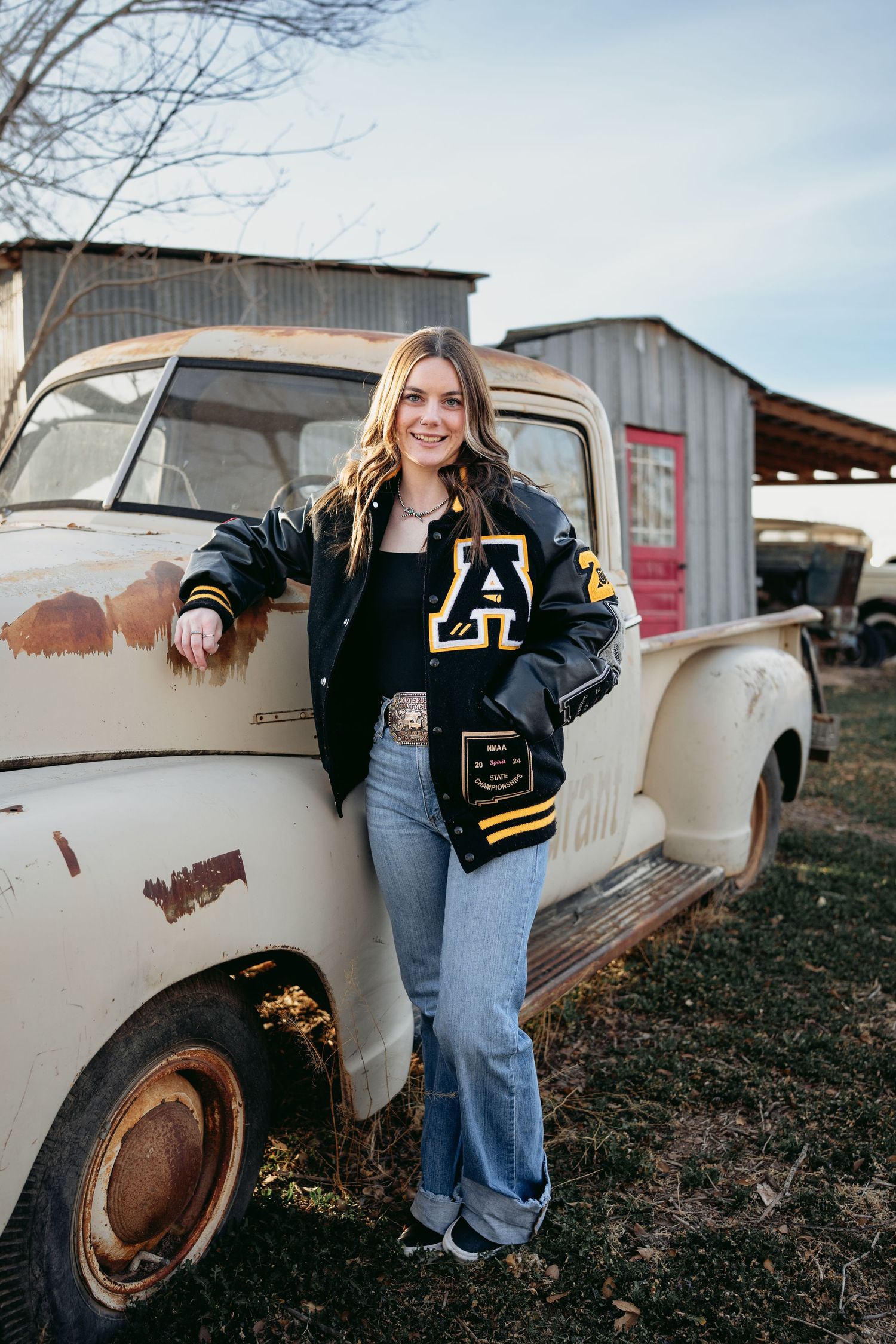 Senior girl in black gold letterman jacket leaning against vintage rusted white pickup truck outdoors.