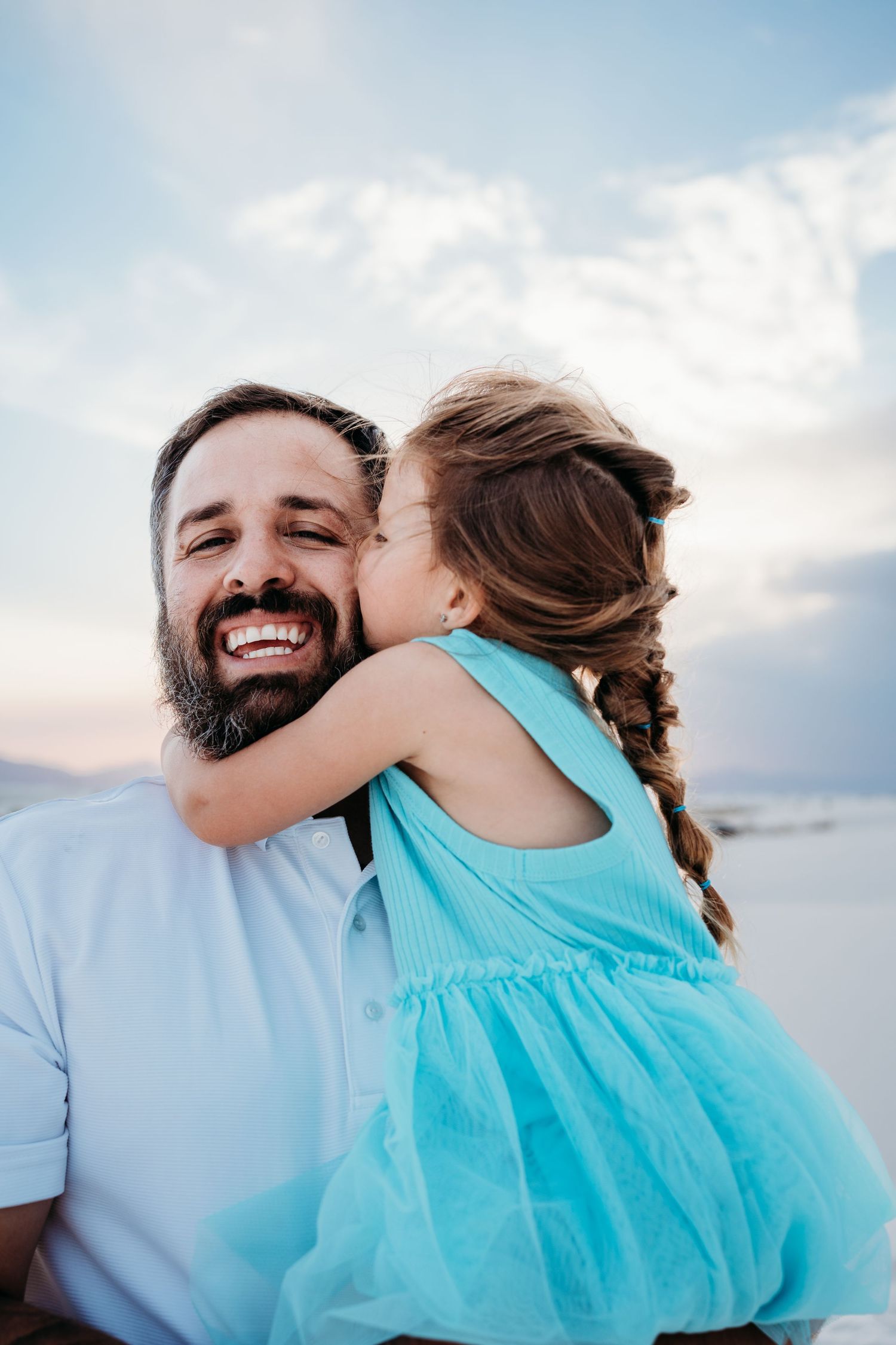 Bearded father smiling joyfully as young daughter in turquoise dress kisses his cheek on sandy beach.