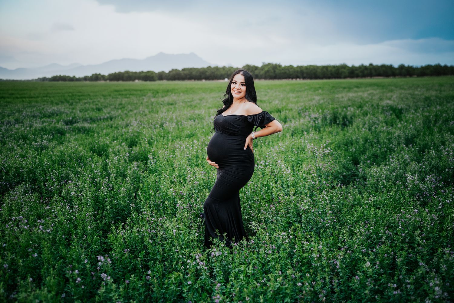 Glowing pregnant woman in a black off-shoulder gown posing in a vibrant green meadow.