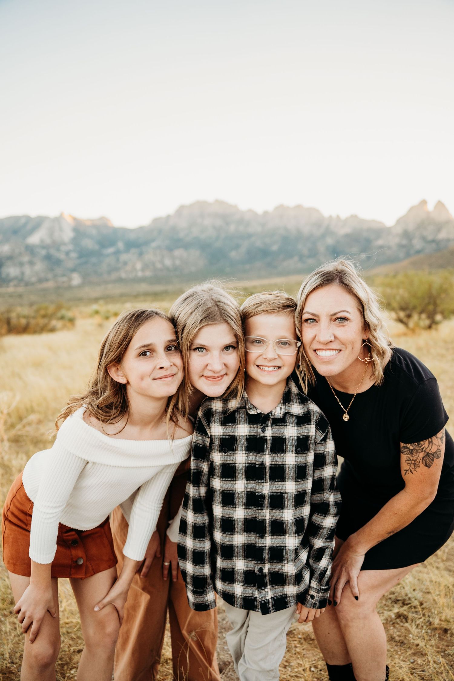 Mother and three children smile together in golden field with dramatic mountain range behind them.