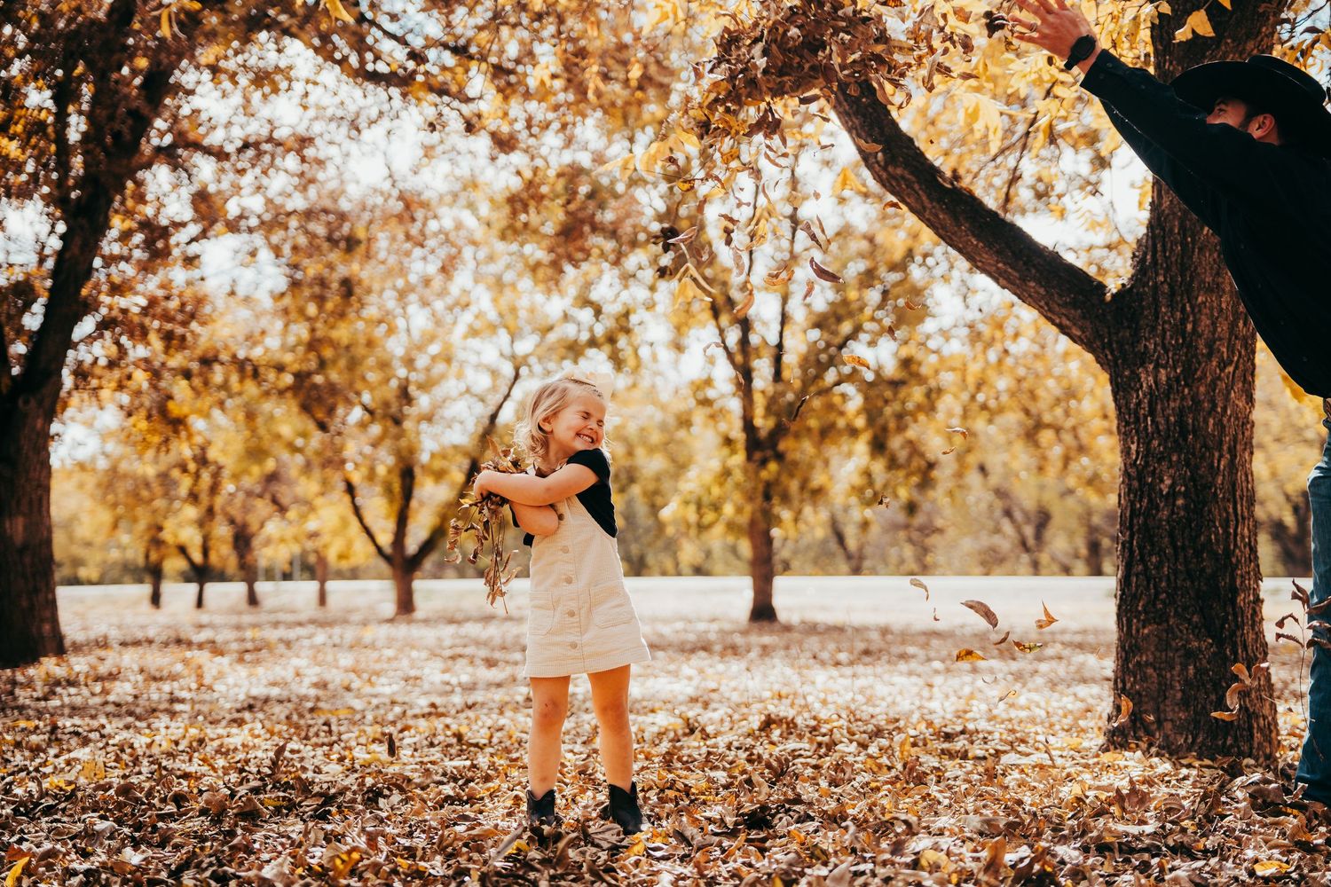 A young girl in a white dress plays among golden fallen leaves in an autumn park.