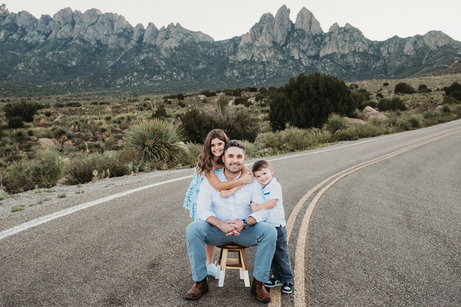 Father seated on stool with pregnant wife and young son posing on mountain road.