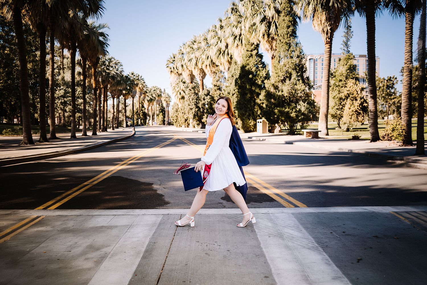 Woman in white dress and navy blazer crossing a palm tree-lined street, carrying a blue bag on a sunny day.