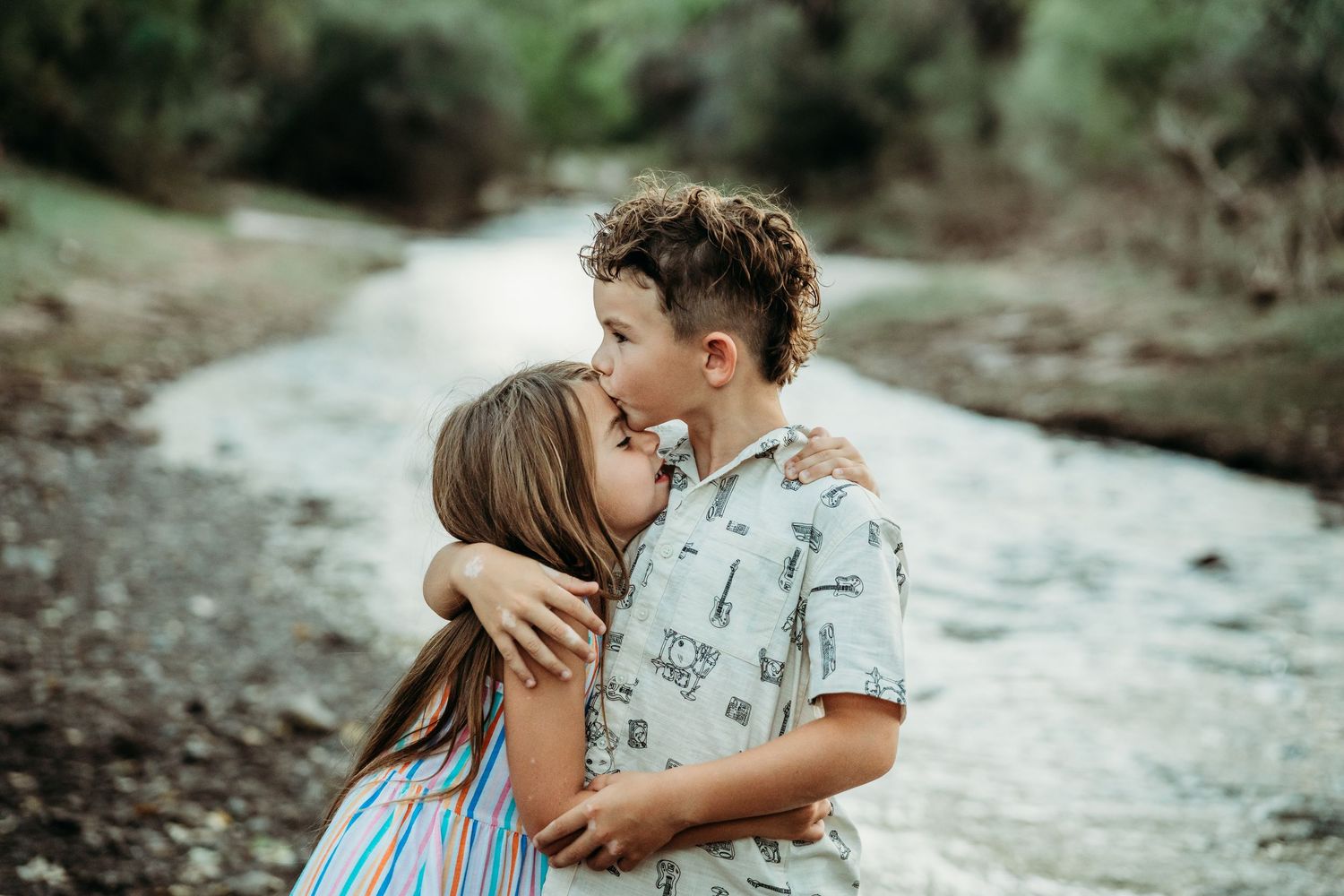 A young girl kissing a boy on the cheek while hugging near a peaceful river outdoors.