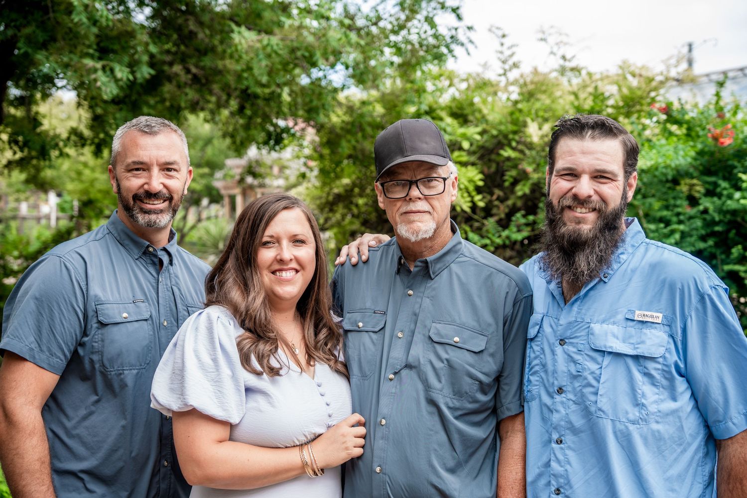 Four men in casual blue shirts smile together outdoors surrounded by lush green trees.